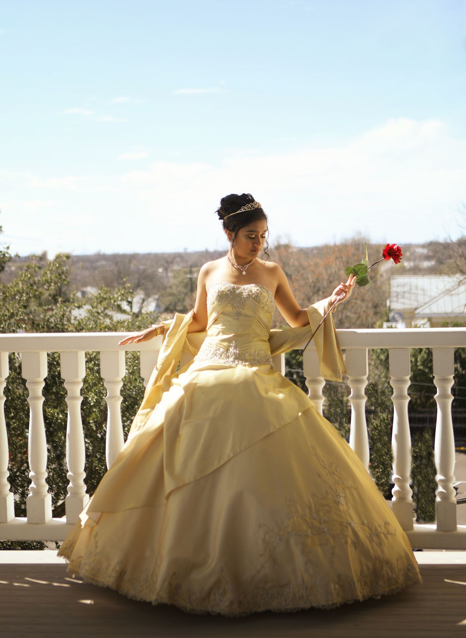 A young girl in a yellow ball gown poses elegantly on a balcony with white railings at the Women's Club in her sweet 16 dress photo session.