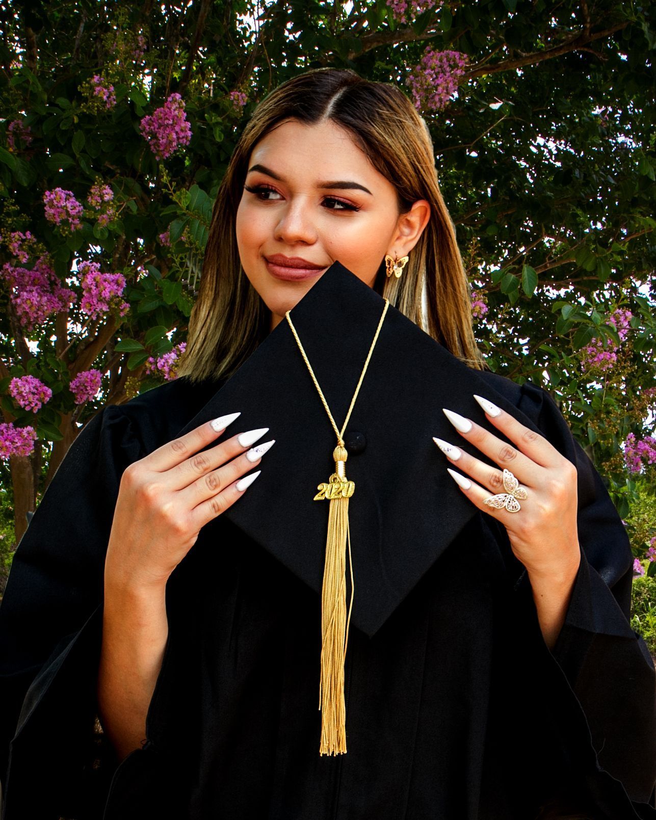 A Graduate in black graduation gown with gold tassel poses in front of blooming pink flowers at Elian Hotal & Spa grounds for senior shoot.