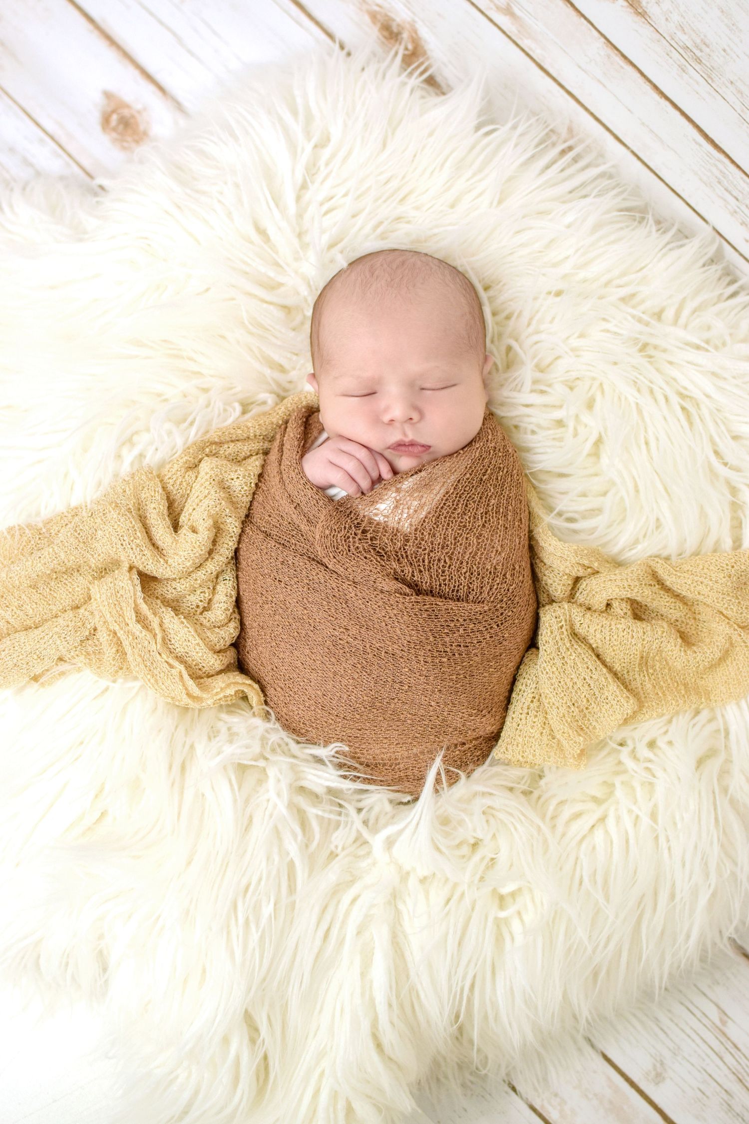 Sleeping newborn baby swaddled in brown wrap on cream fluffy rug for professional newborn photography.