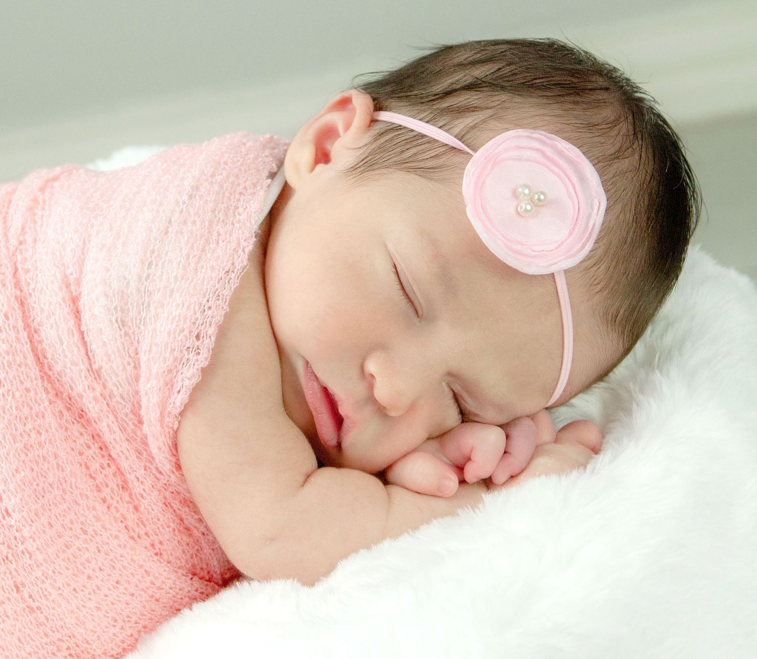 Sleeping newborn baby girl wrapped in pink swaddle with floral headband resting peacefully on white fluffy blanket.