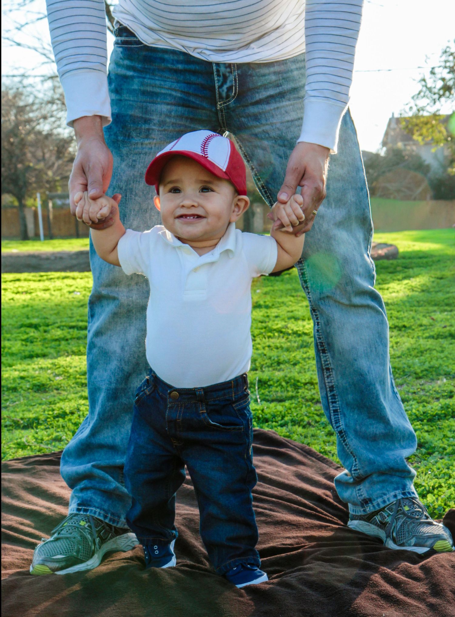 A smiling baby in a red cap and white onesie takes first steps while being supported by an adult outdoors.