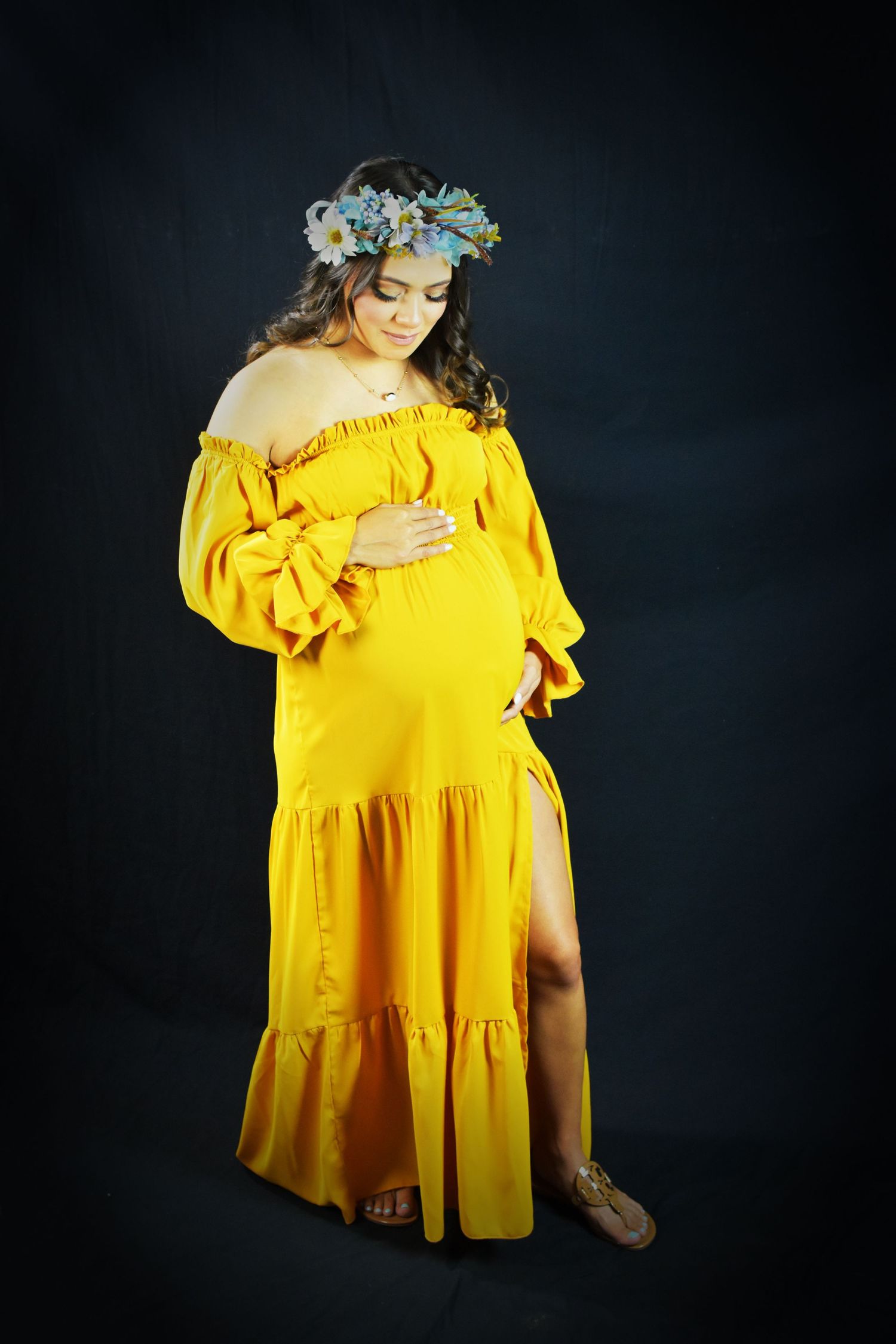 A pregnant woman in a flowing yellow off-shoulder dress and blue floral crown poses against a dark backdrop at her luxury maternity session.