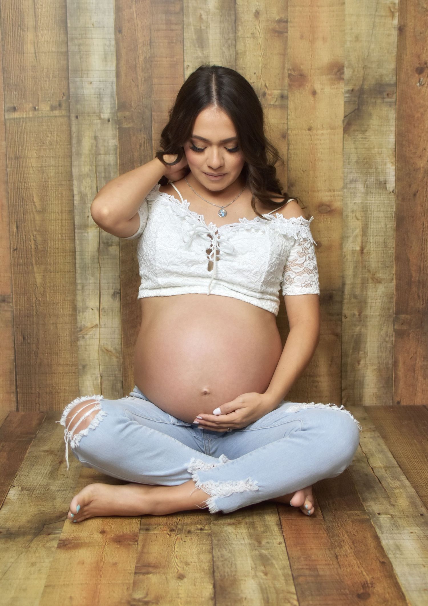A maternity photo shows a pregnant woman in white lace top and jeans sitting on wooden floor in San Antonio, Texas