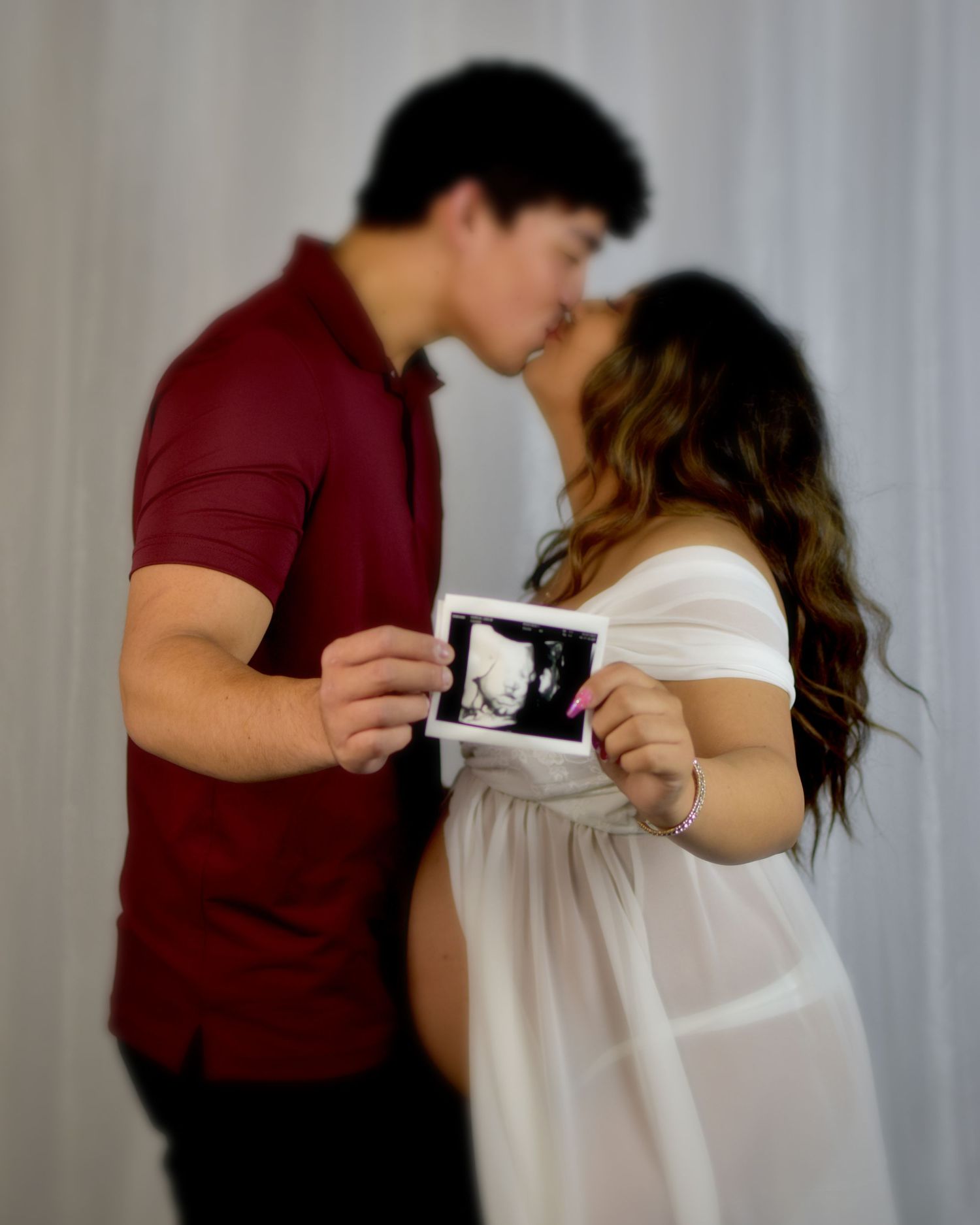A couple in burgundy and white clothing share an intimate kiss holding their baby in sonogram in maternity photo session in San Antonio, Tx