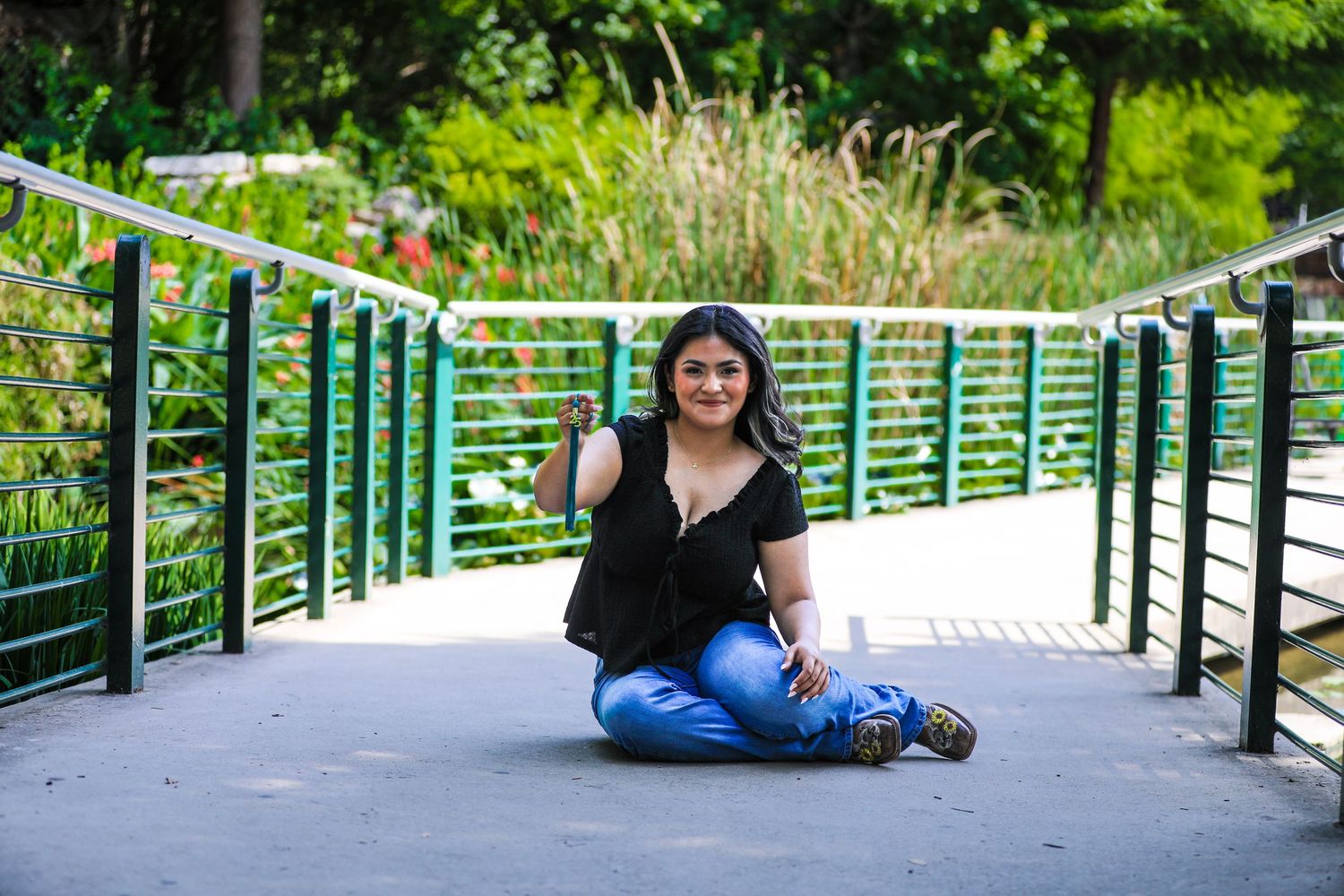 A student in blue jeans sits on a bridge at The Pearl showing her tassel at her senior photography session in San Antonio, Texas