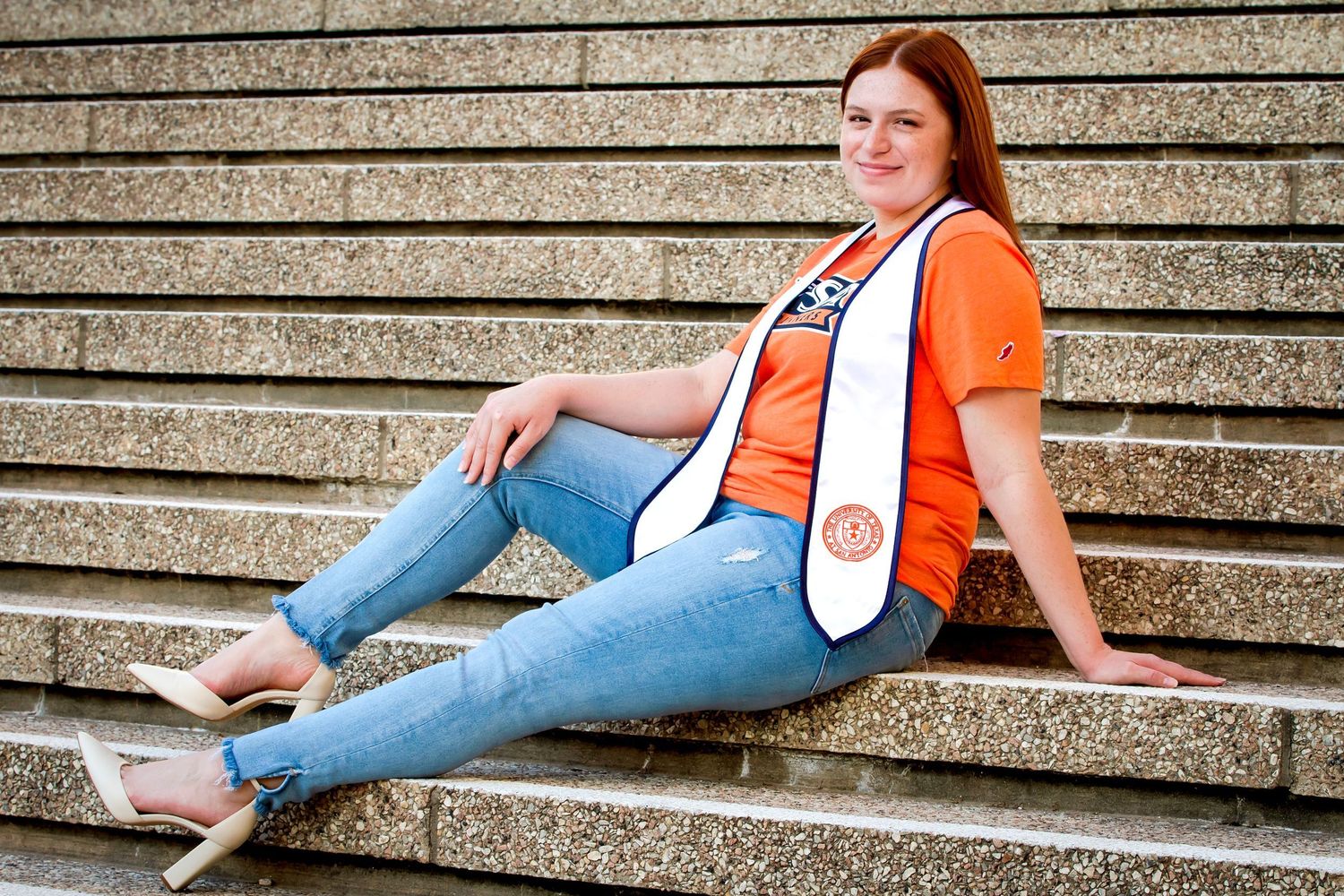 Student in orange UTSA t-shirt lounges casually on concrete steps during her senior/graduation photography session in San Antonio, Texas