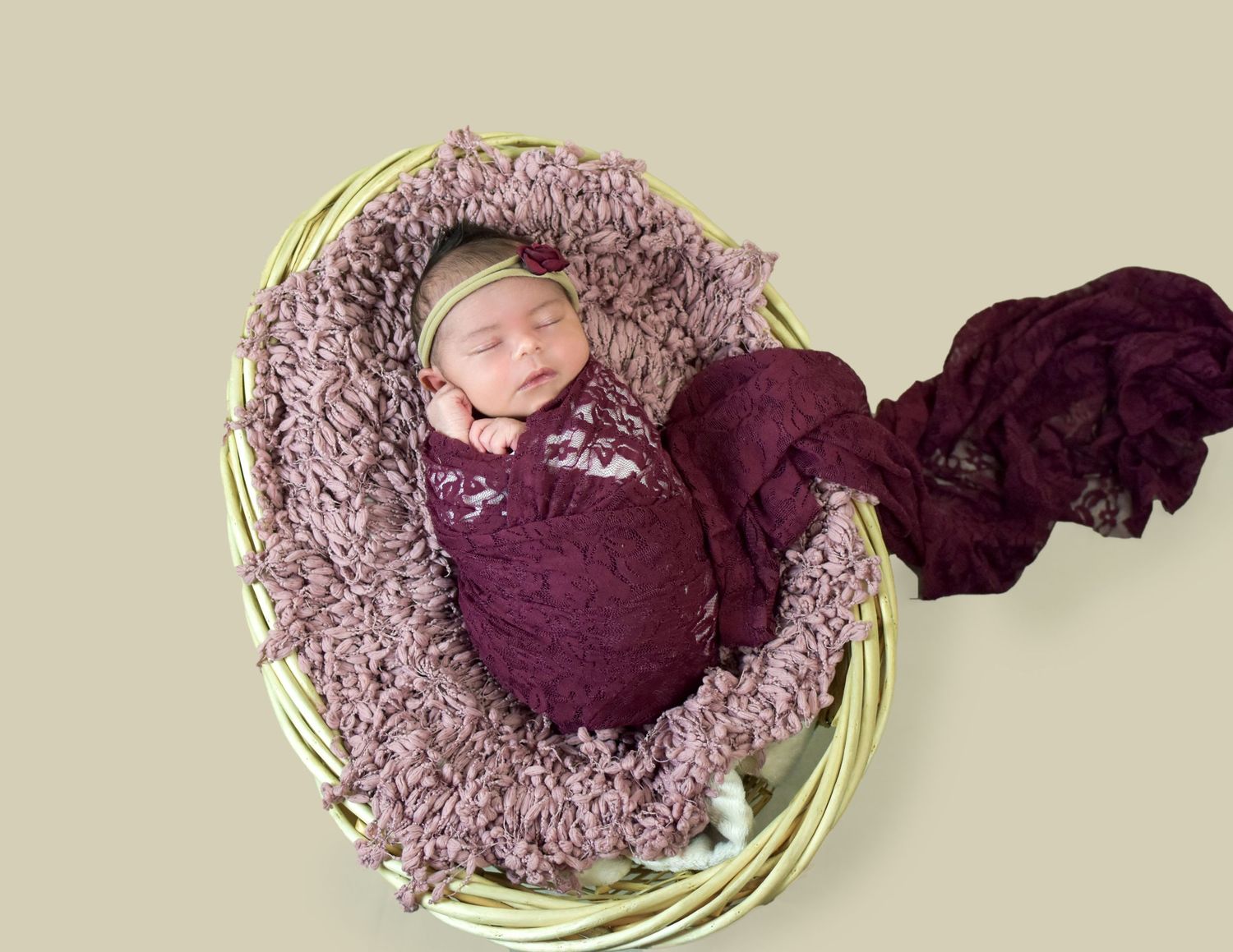 Newborn baby sleeping in a wicker basket wrapped in purple lace with a matching floral headband.