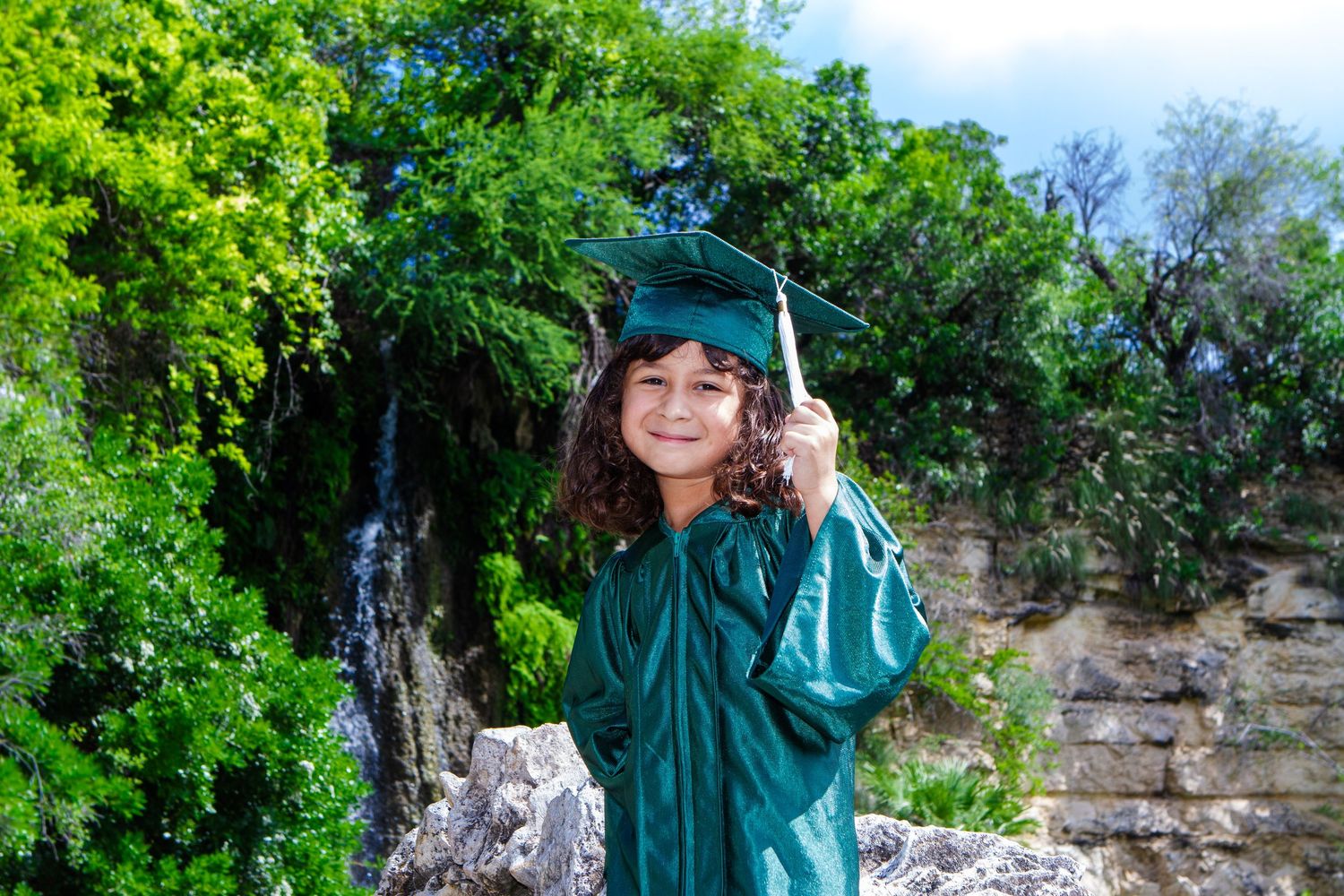 Young child in green graduation cap and gown smiling outdoors near a waterfall and lush green trees.