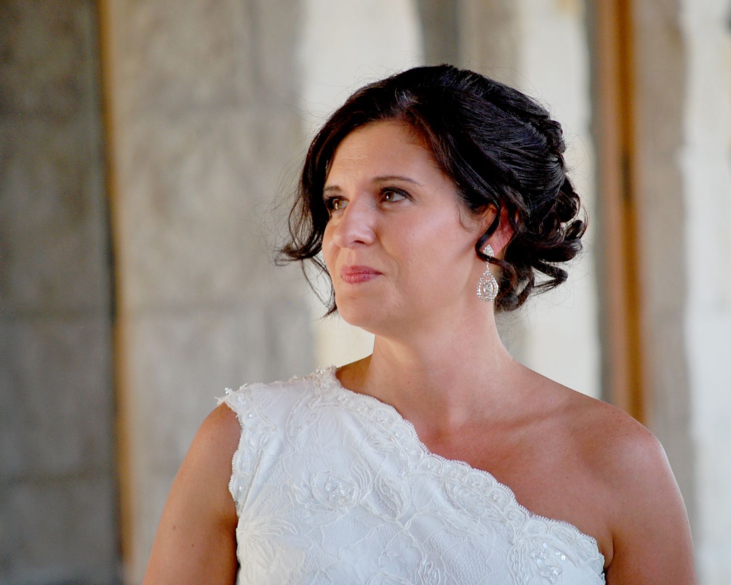 A bride in a white one-shoulder gown with an elegant updo hairstyle and pearl earrings gazes thoughtfully.