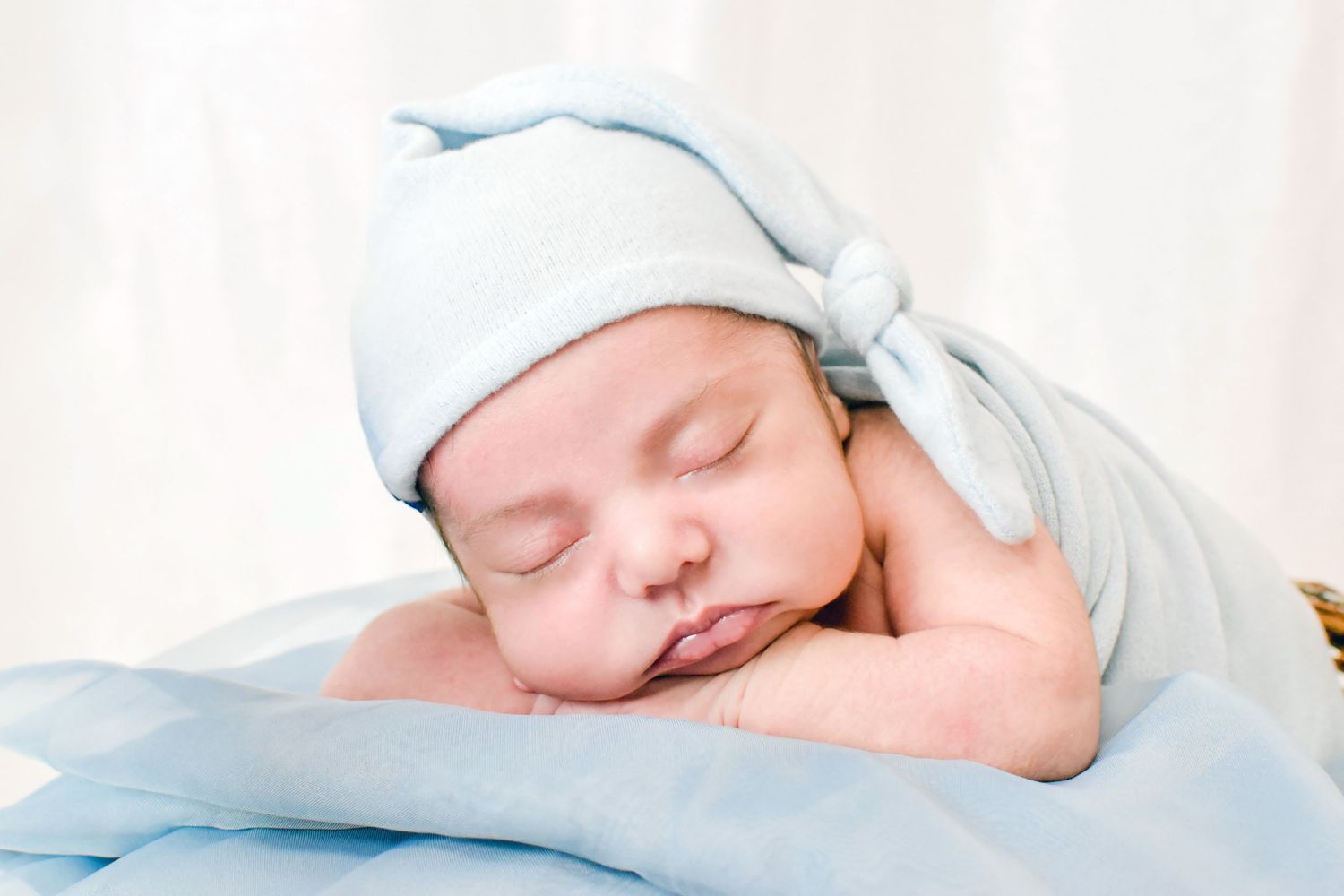Peaceful newborn baby sleeping in a blue knit hat and wrap, resting on a soft blue blanket.