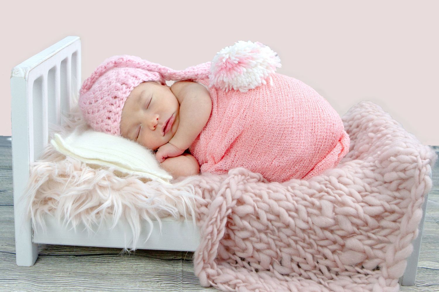 Newborn baby sleeping peacefully in a tiny white bed, wearing a pink knit outfit and bunny hat.
