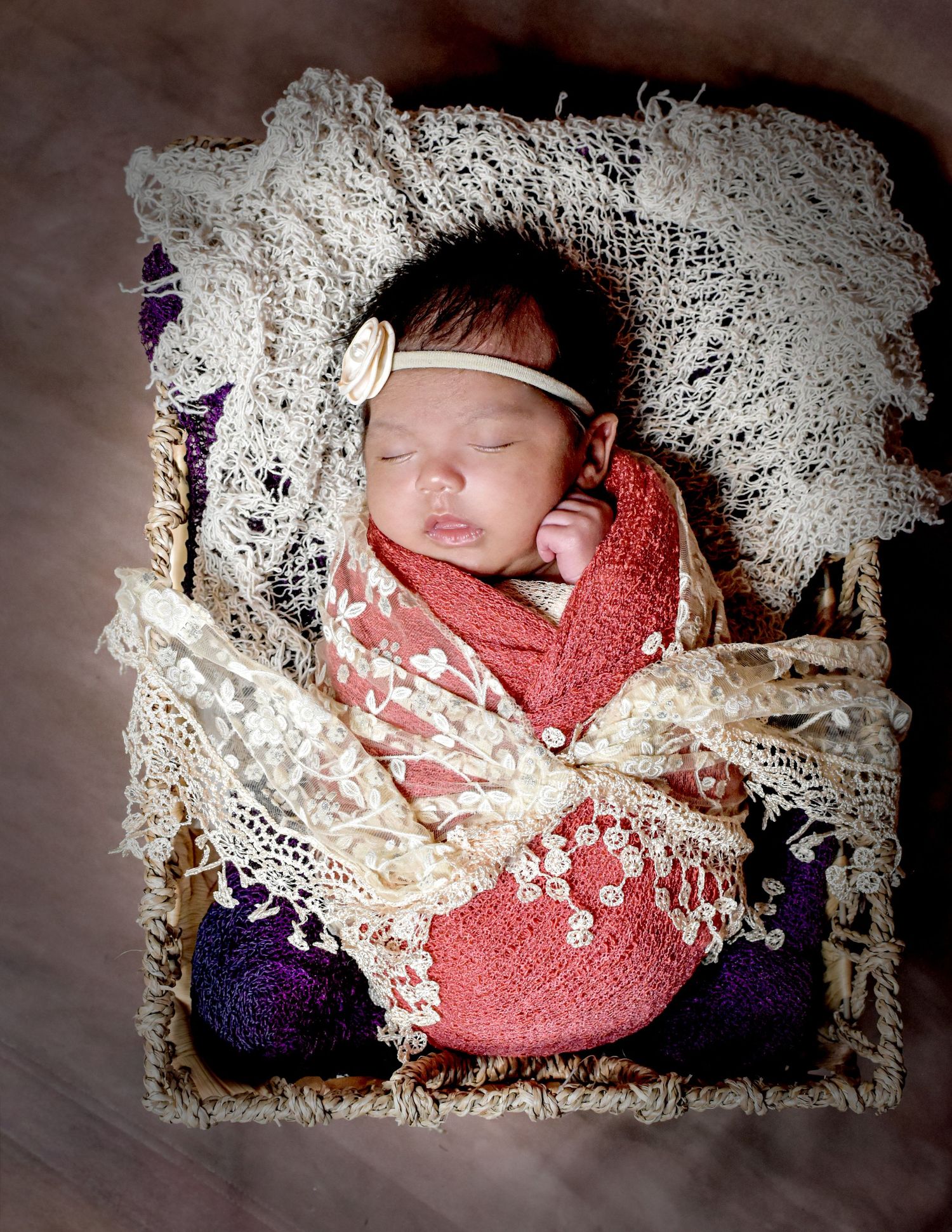Newborn baby sleeping peacefully wrapped in pink lace swaddle with floral headband in rustic basket.