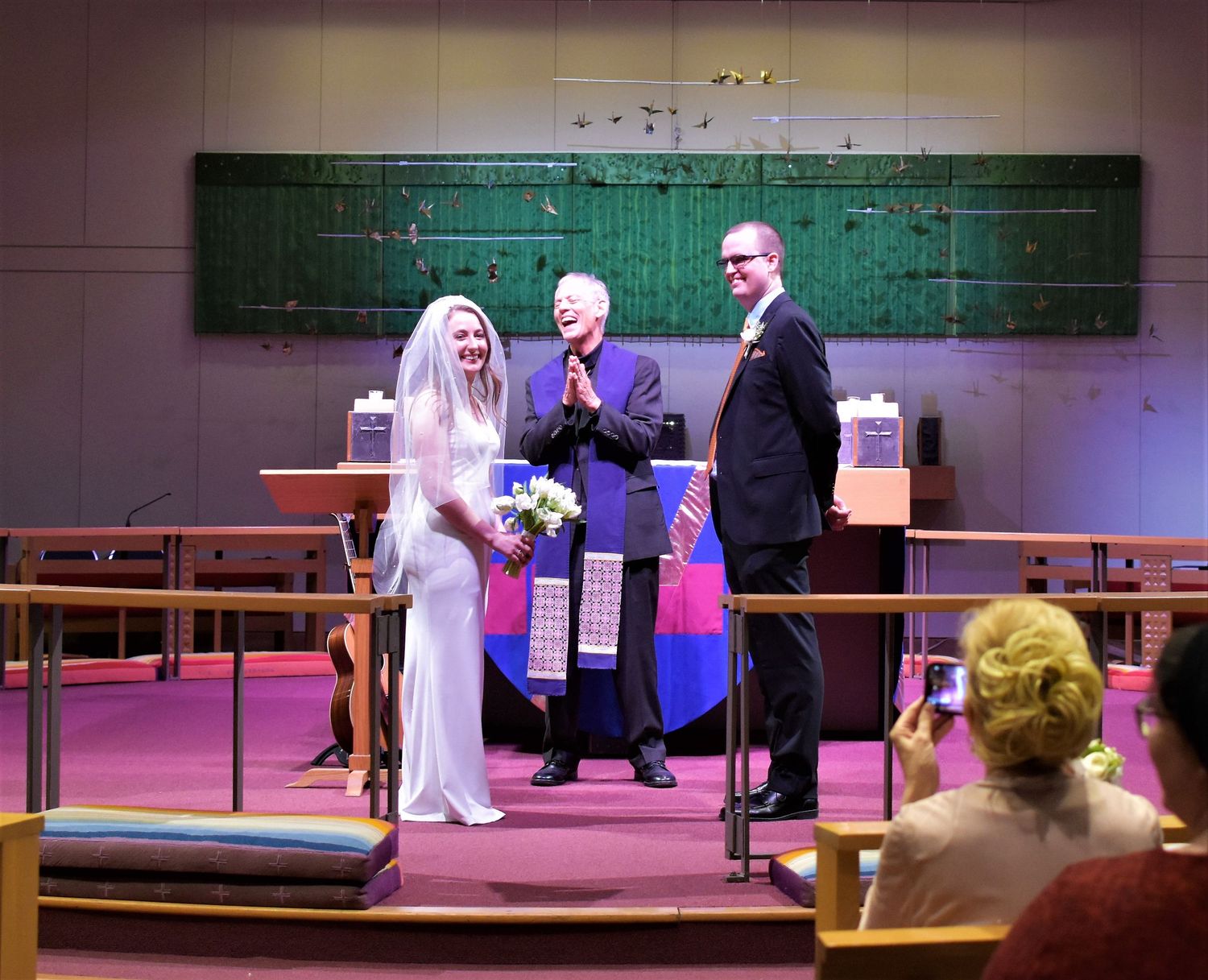 A bride and groom stand with an officiant during a wedding ceremony inside a church with pink carpeting.