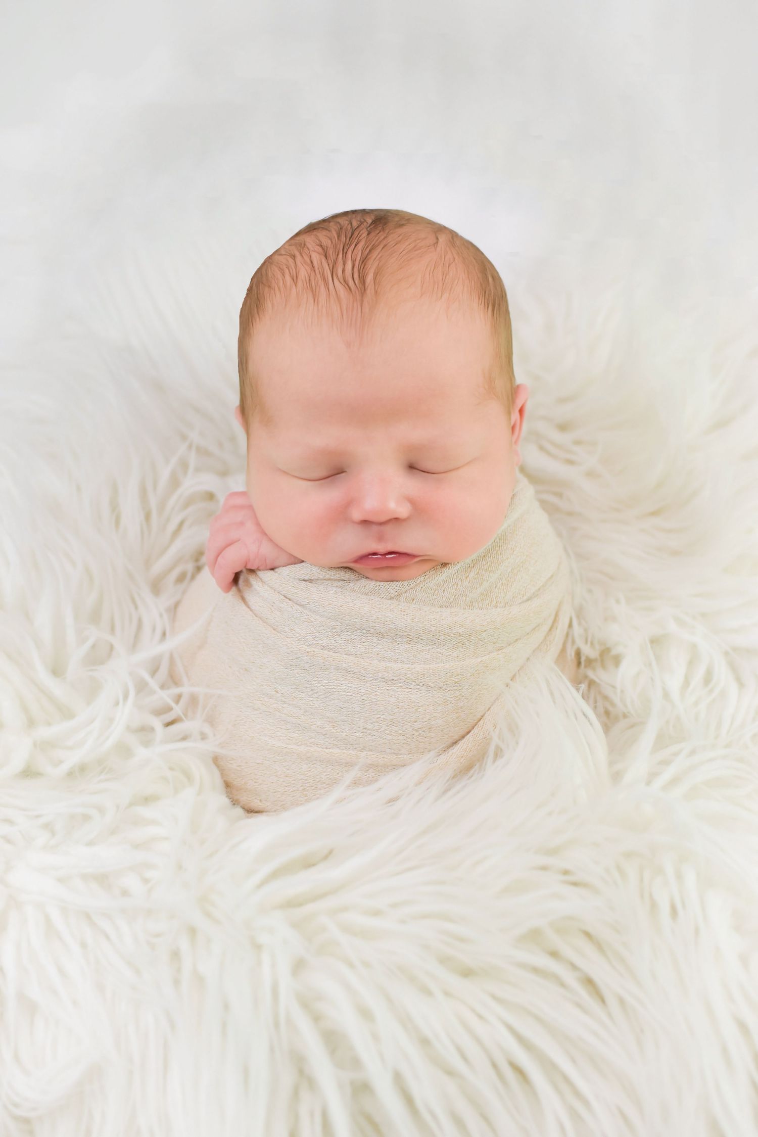 Sleeping newborn baby swaddled in cream fabric resting on soft white fur background.