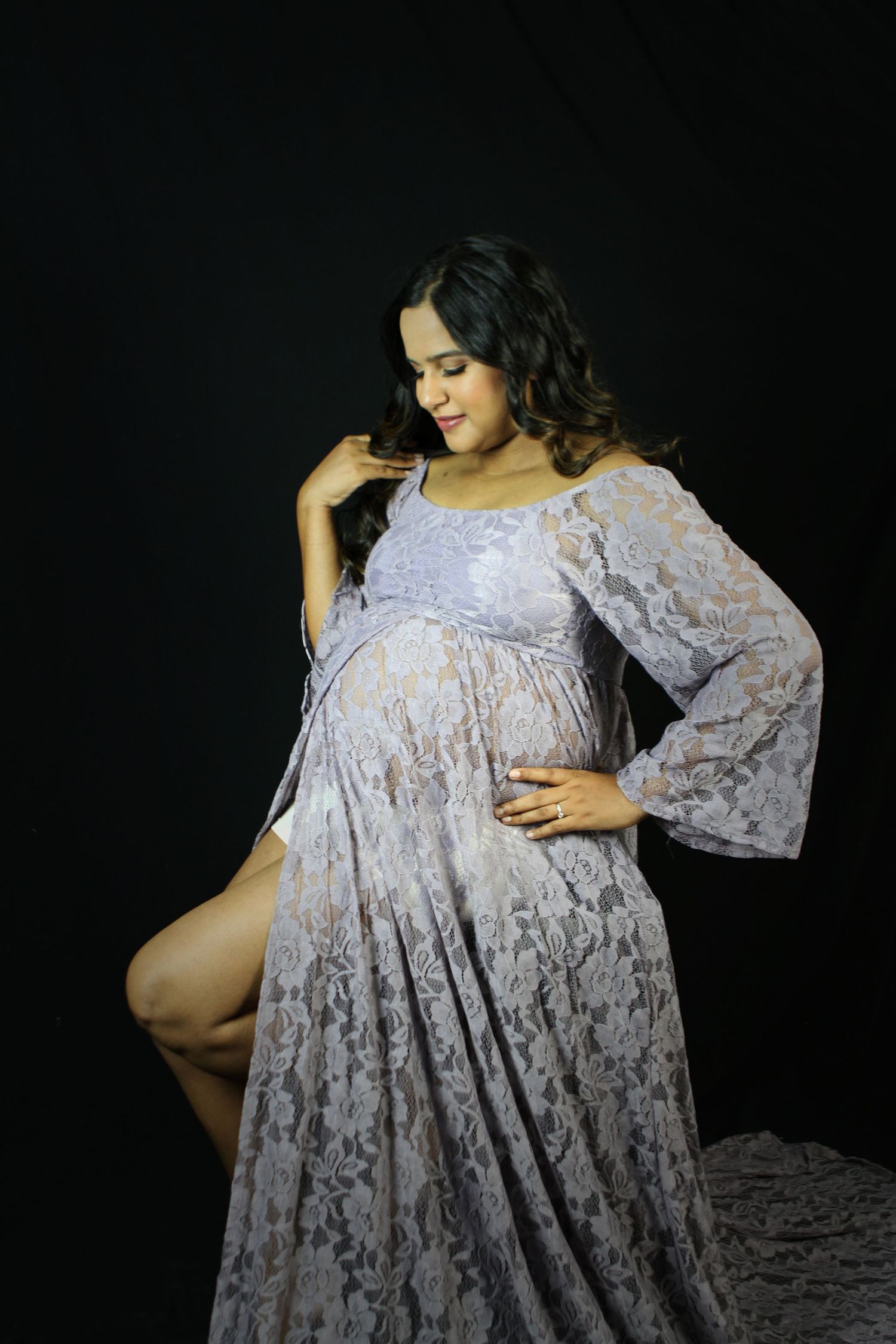 Pregnant woman in elegant lavender lace maternity gown posing gracefully against black studio background.
