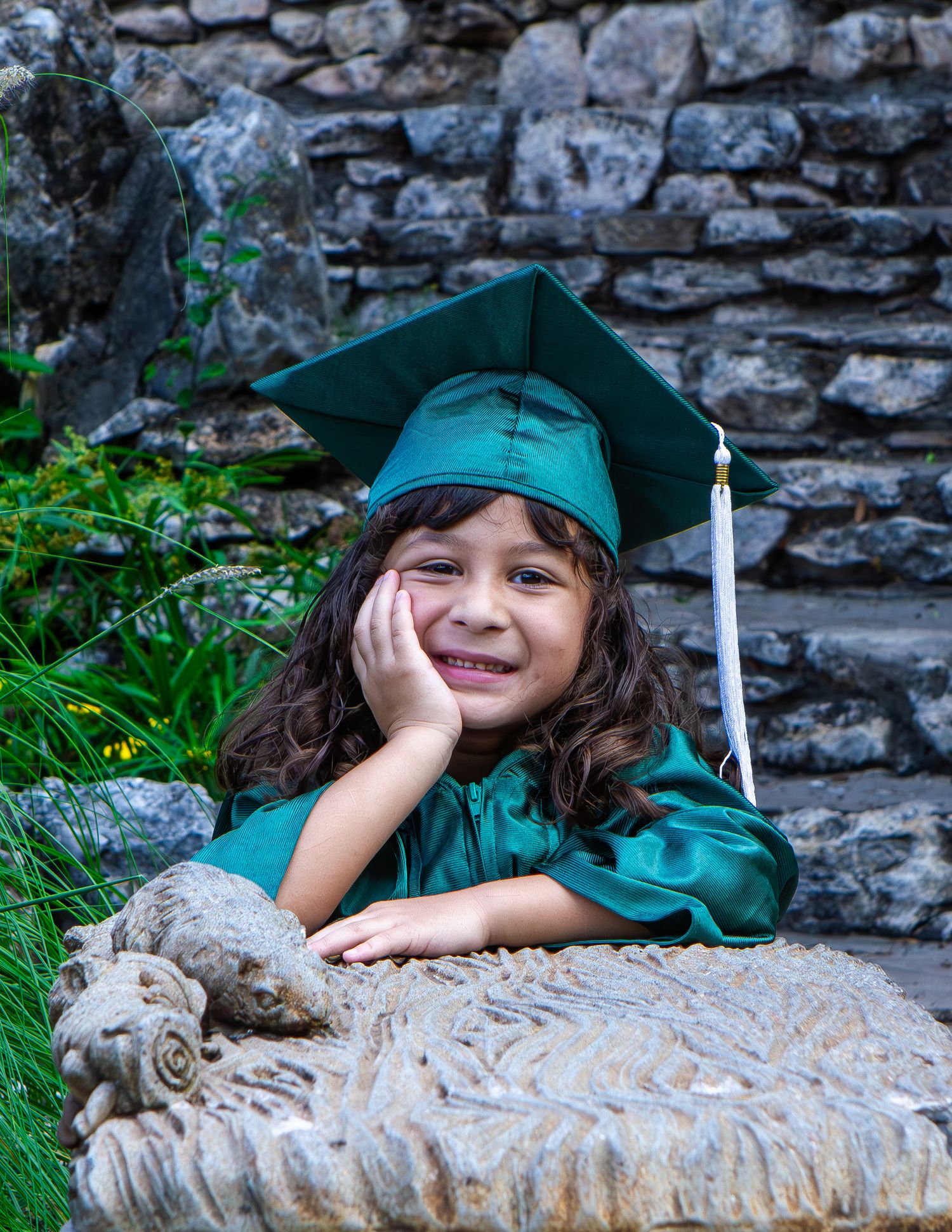 Young girl in green graduation cap and gown smiling while leaning on stone bench outdoors.