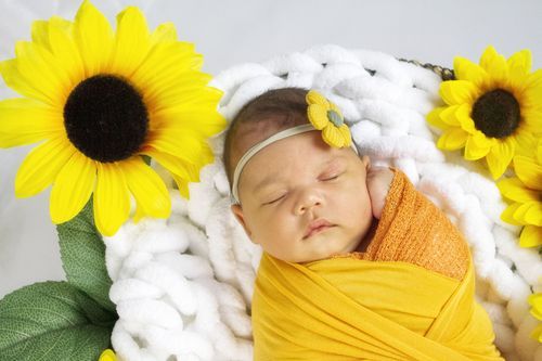 Newborn baby swaddled in yellow wrap sleeping peacefully on white chunky knit blanket surrounded by bright sunflowers.