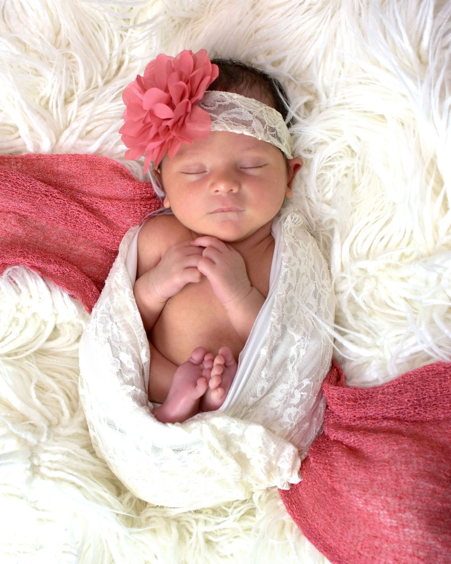 Sleeping newborn baby girl wrapped in white lace and pink swaddle, wearing a coral flower headband on fluffy white rug.