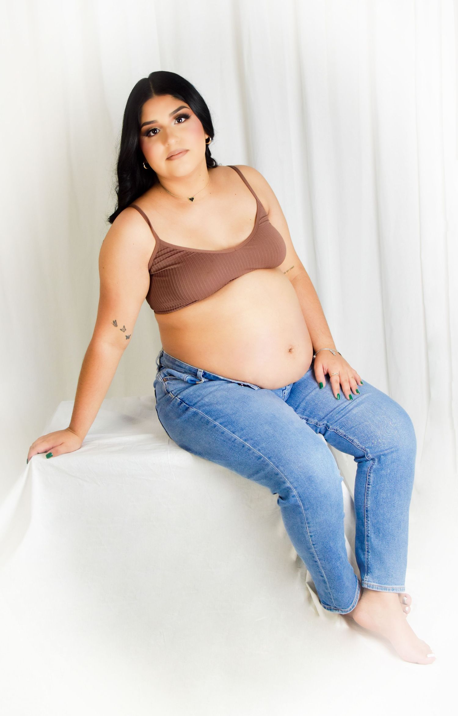 A maternity photo session mom to be wearing a brown crop top and blue jeans sits against a white backdrop in a casual maternity pose. luxury