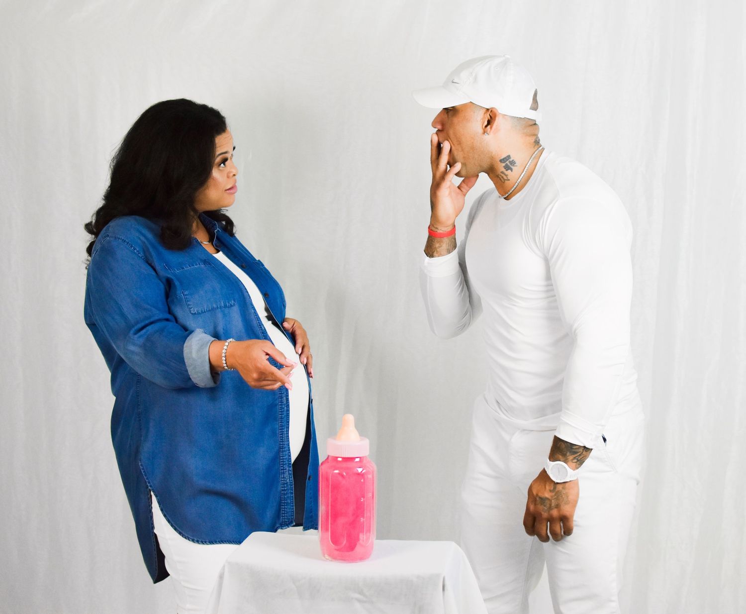 Pregnant woman in blue denim jacket stands with man in white outfit near pink baby bottle at gender reveal. Precious Keepsakes Photography
