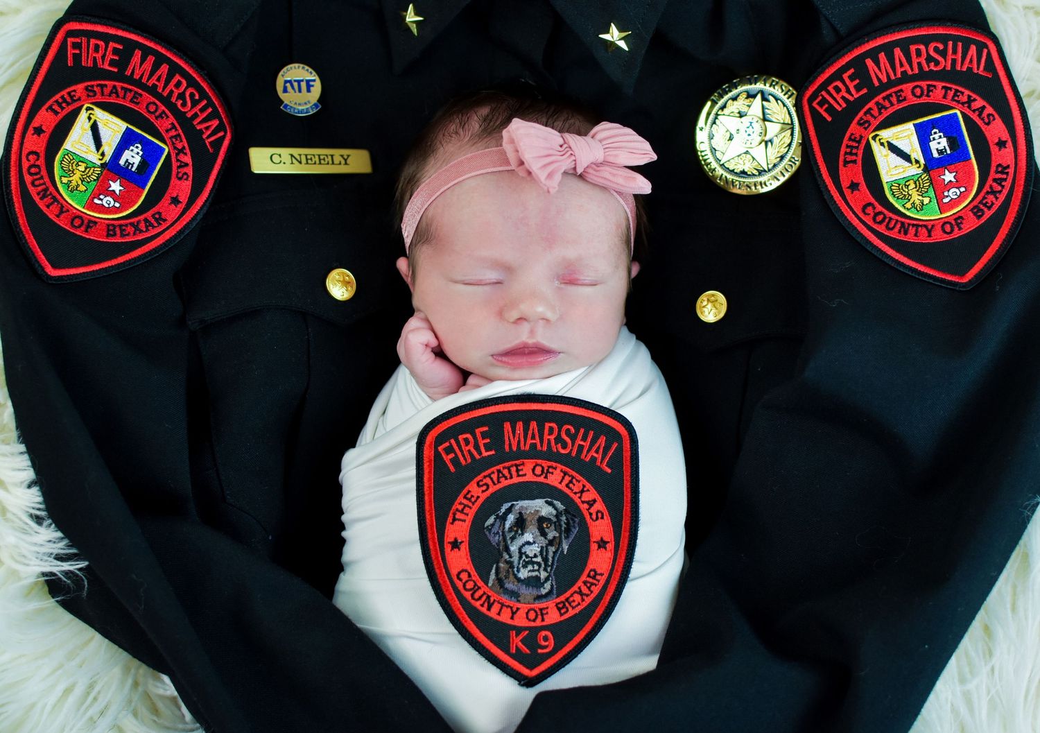 Newborn baby sleeping on Fire Marshal uniform with patches and badges, wearing white onesie and pink bow.