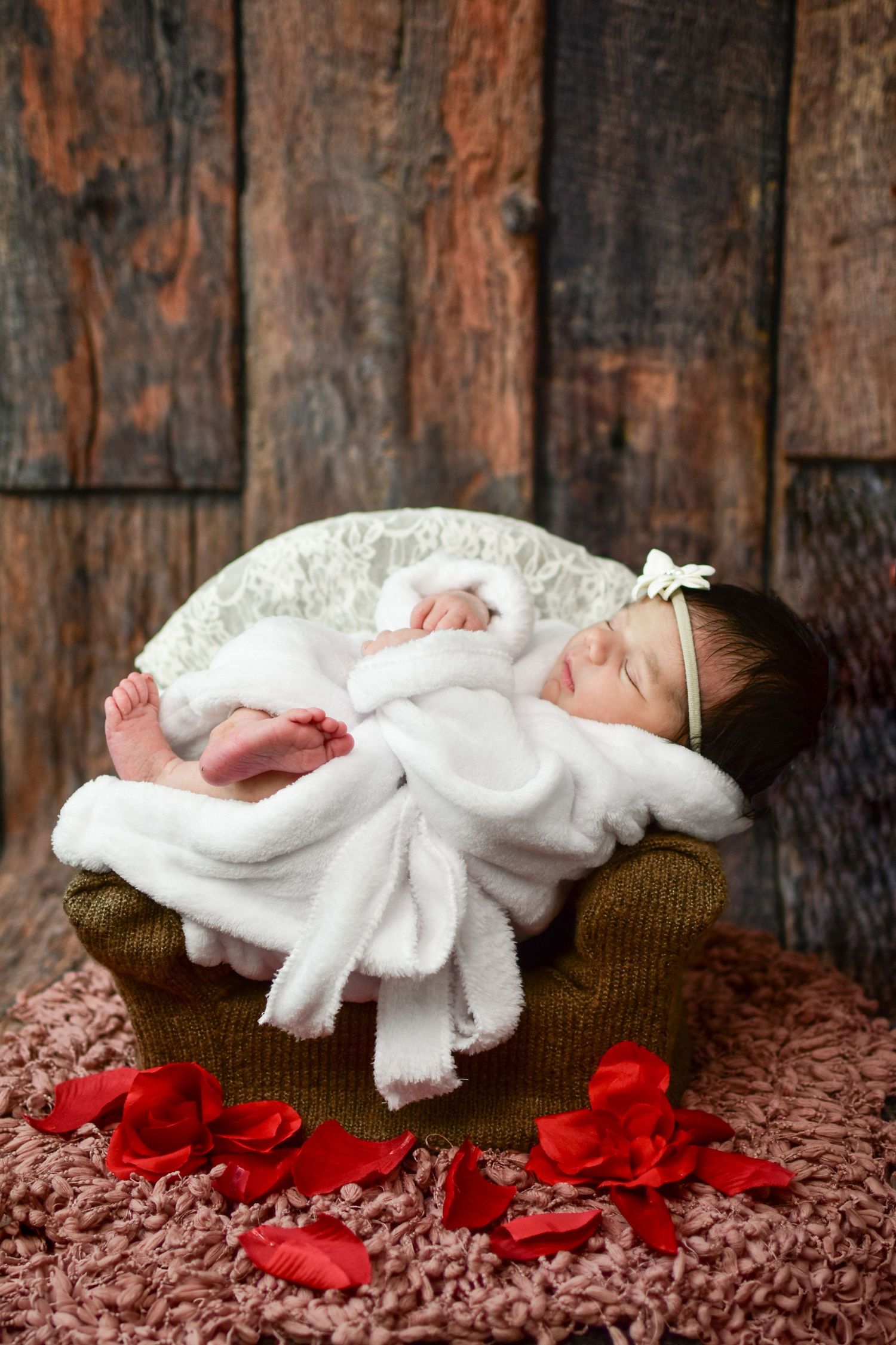 Newborn baby girl sleeping peacefully in a tiny chair, wrapped in white, with red rose petals on pink rug.