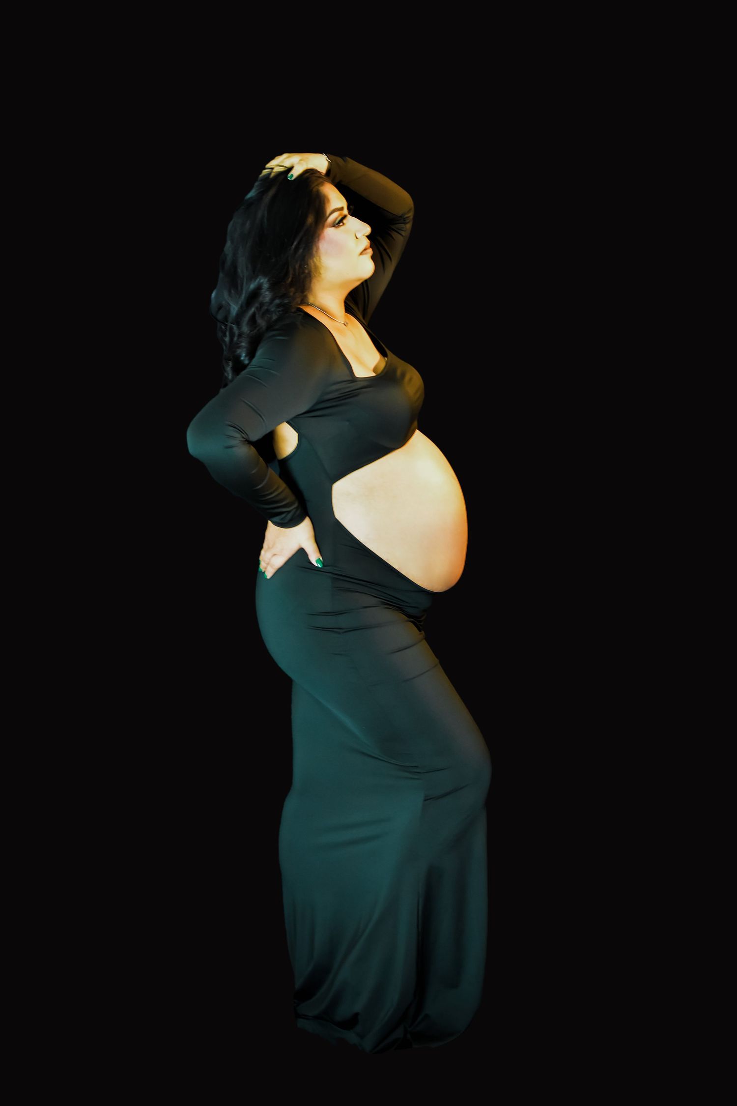 A dramatic silhouette shows a mom to be in black dress showing her amazing baby bump curves against a dark background in San Antonio, Texas