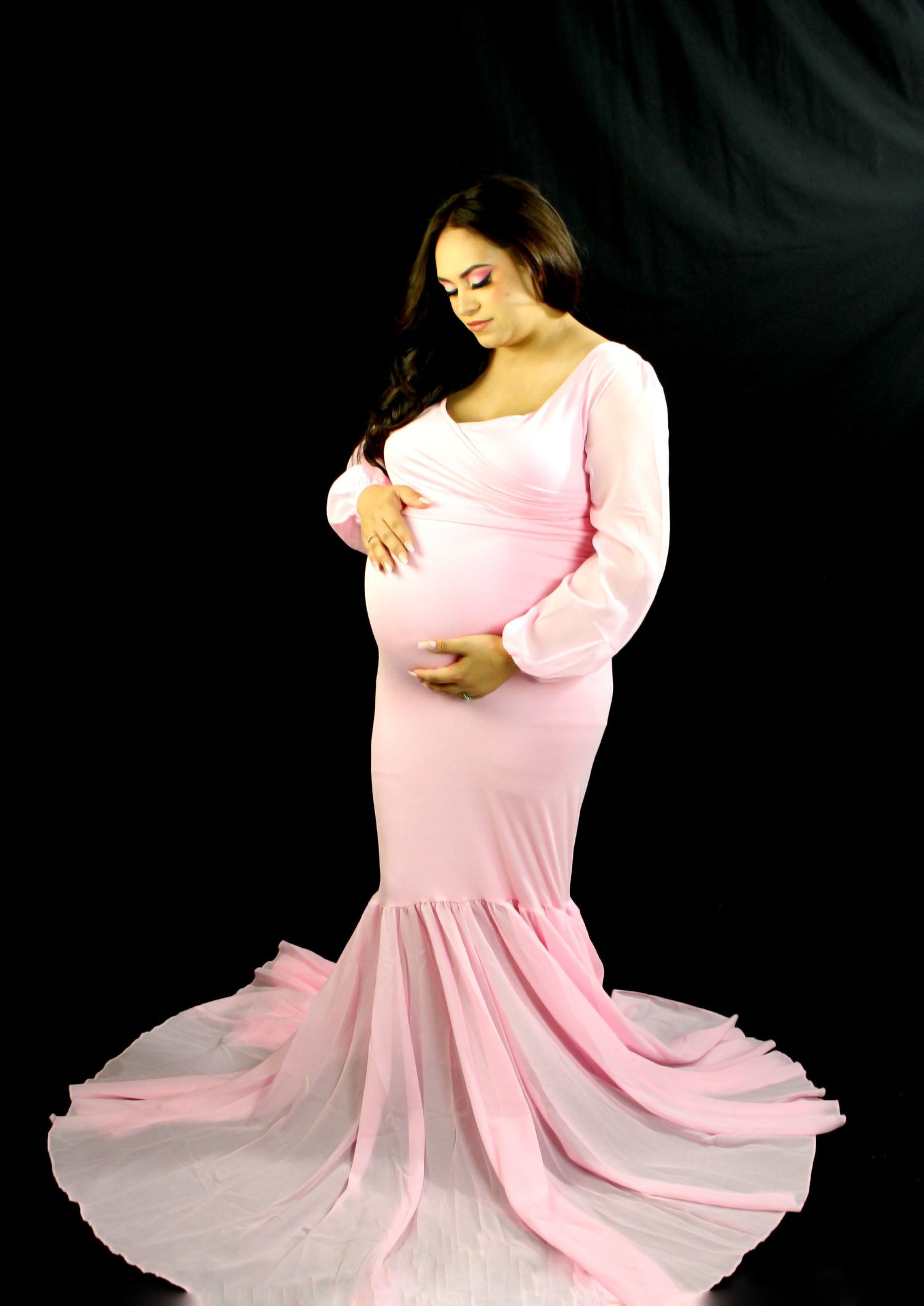 A pregnant woman in a flowing pink maternity gown and cardigan poses gracefully against a dark background holding her baby bump San Marcus