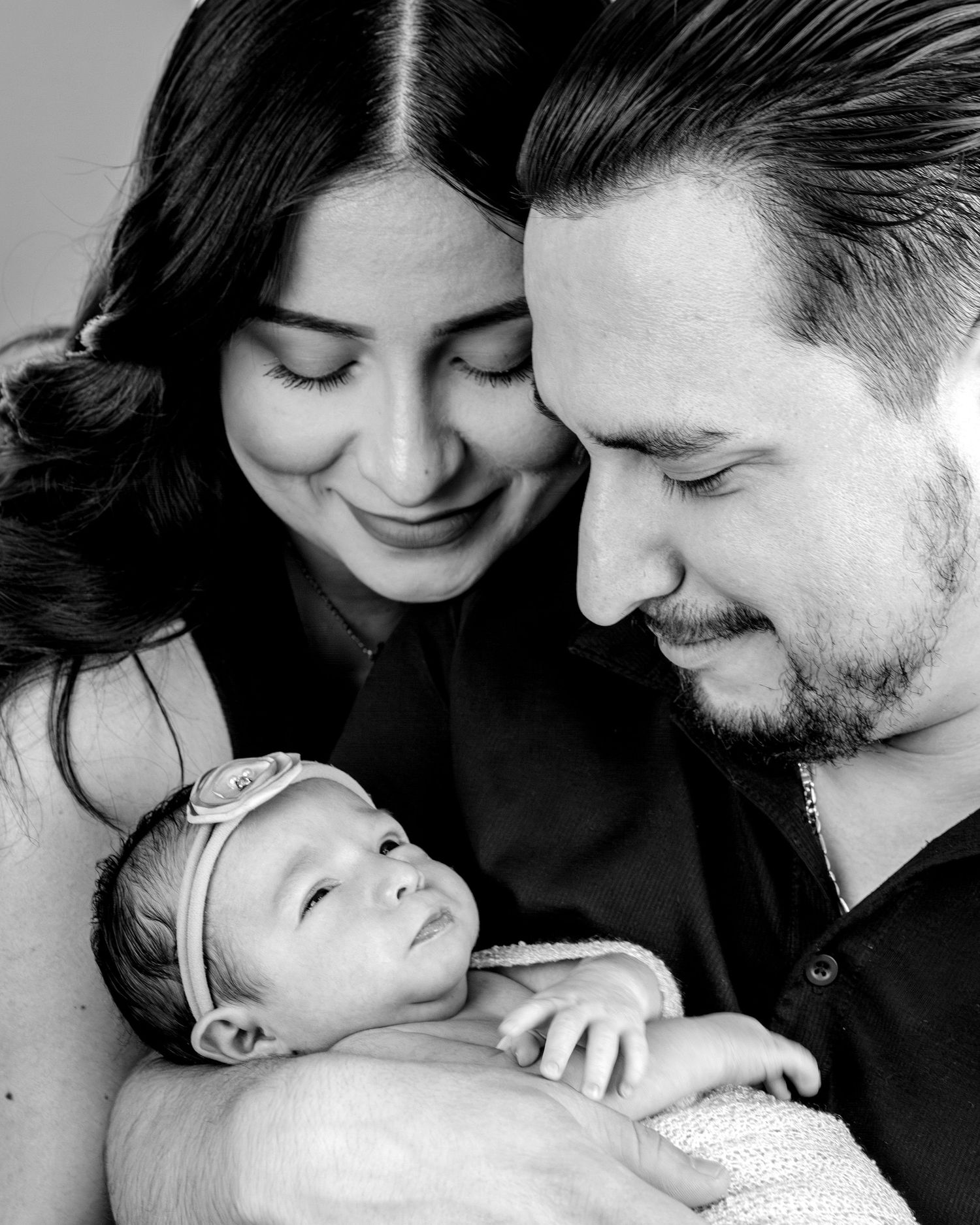 Black and white photo of smiling parents lovingly gazing at their newborn baby girl wearing a floral headband.