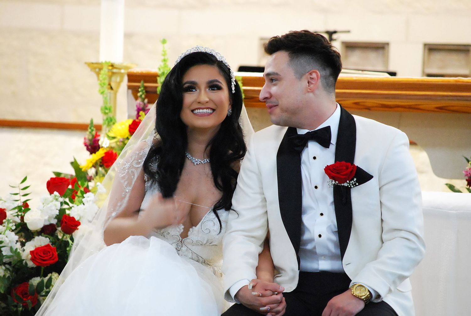 Happy couple in wedding attire smiling at church altar, bride in white gown and groom in white tuxedo with red rose.