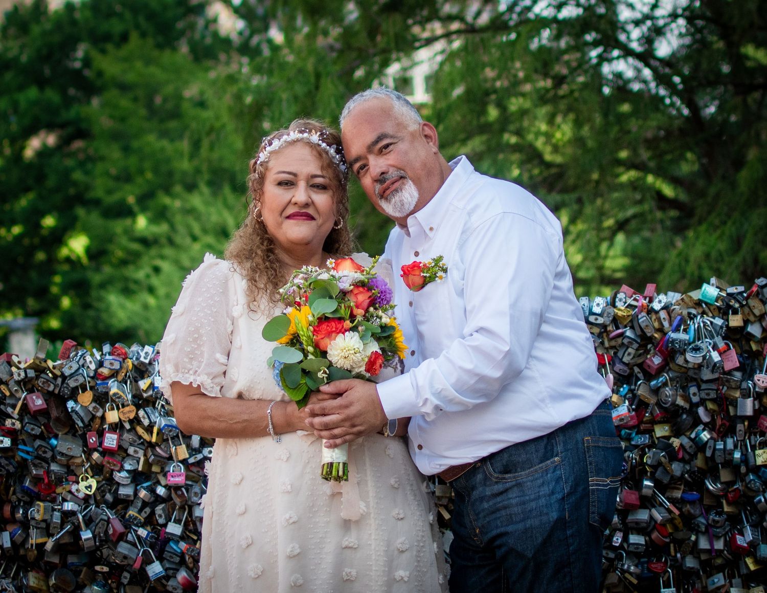 A couple in wedding attire poses smiling before a colorful love lock fence, bride holding a vibrant bouquet.