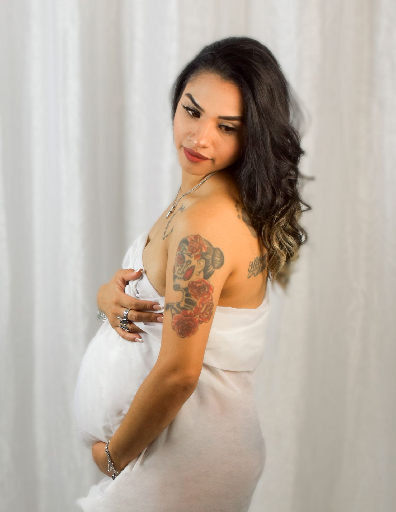 Pregnant woman in white wrap dress showing colorful floral tattoo on shoulder, posing sideways in studio. Precious Keepsakes Photography