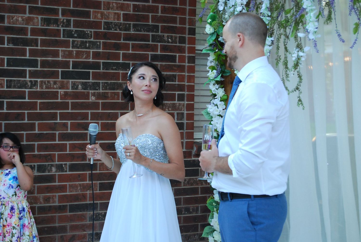 Bride in white gown and groom in blue pants share a moment during outdoor wedding ceremony by brick wall.