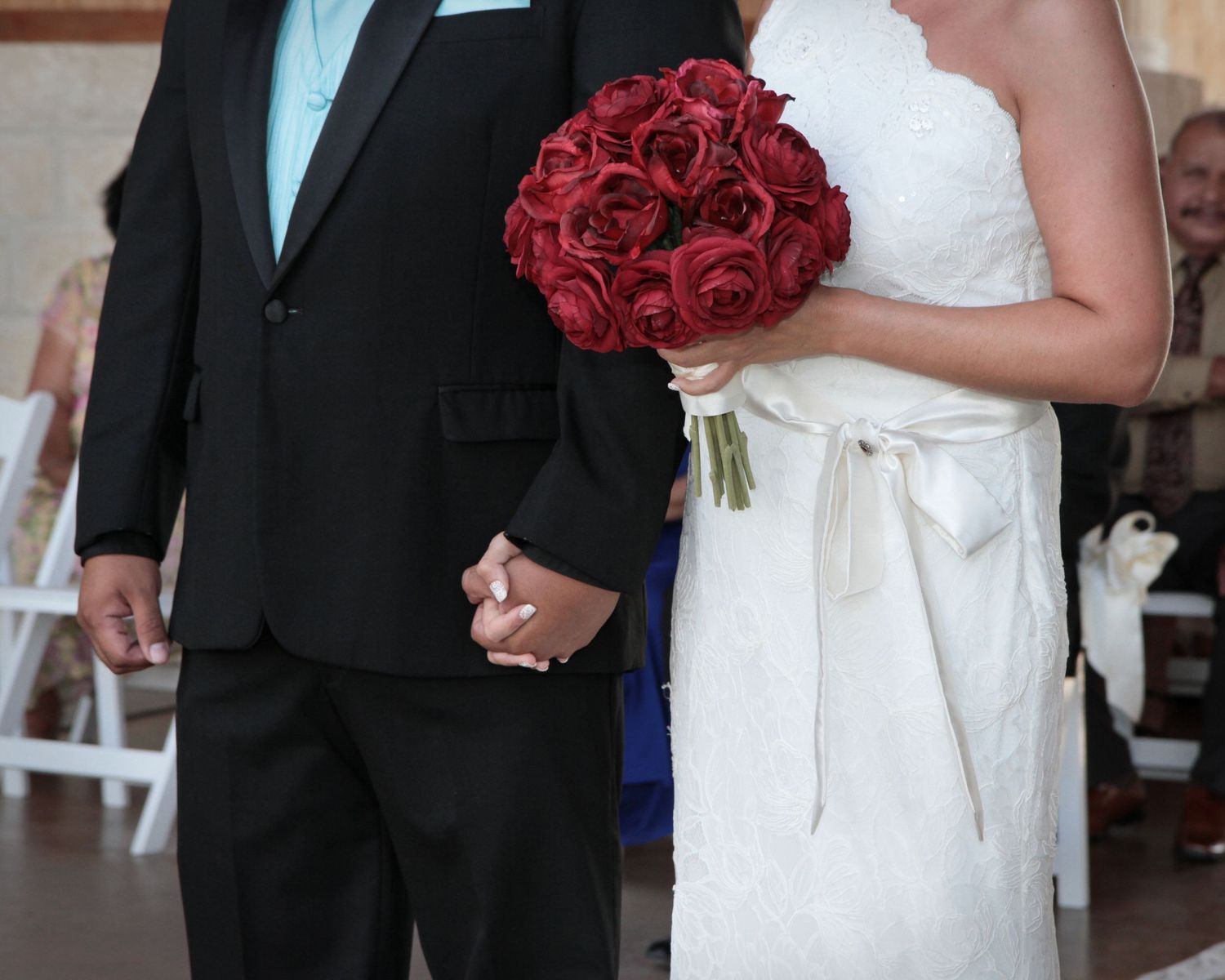 Bride in lace gown holding red rose bouquet stands with groom in black tuxedo at wedding ceremony.