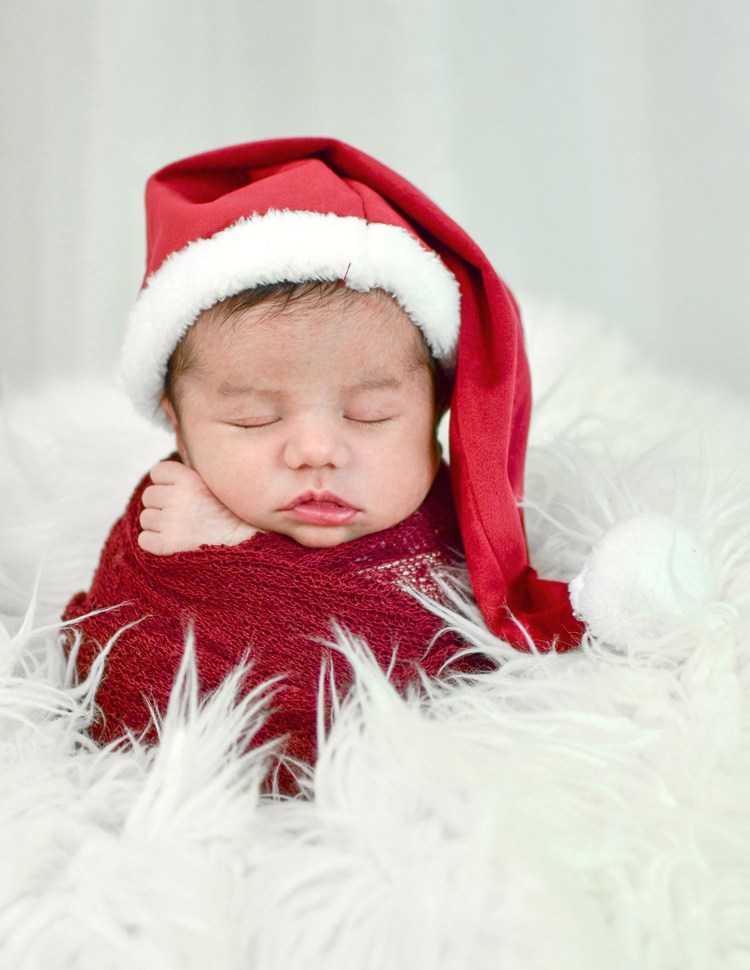 Newborn baby sleeping peacefully in a red Santa hat and wrap on white fluffy fur, perfect Christmas holiday photo.