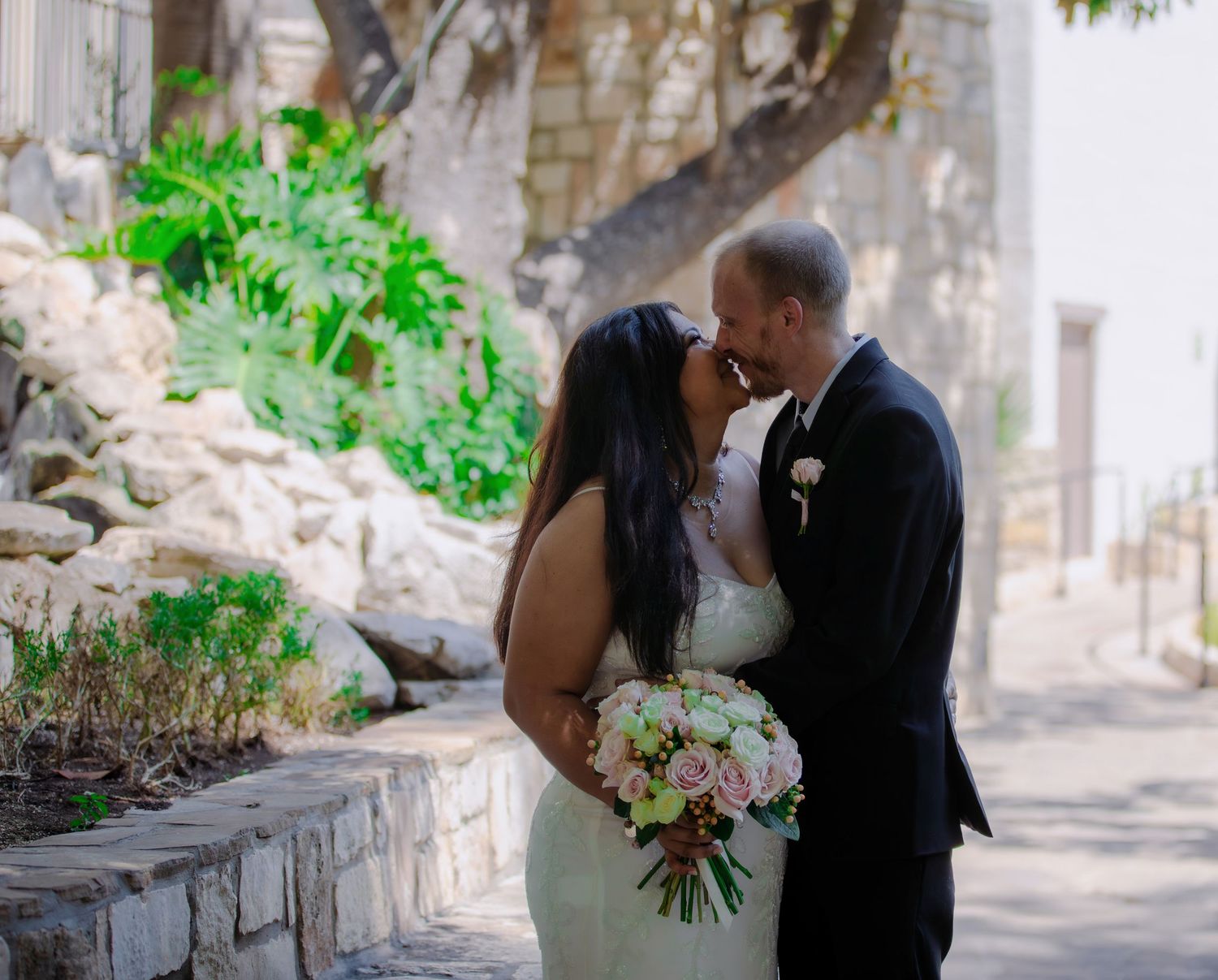 A bride and groom share a romantic kiss outdoors near stone walls, bride holding a pink and white bouquet.