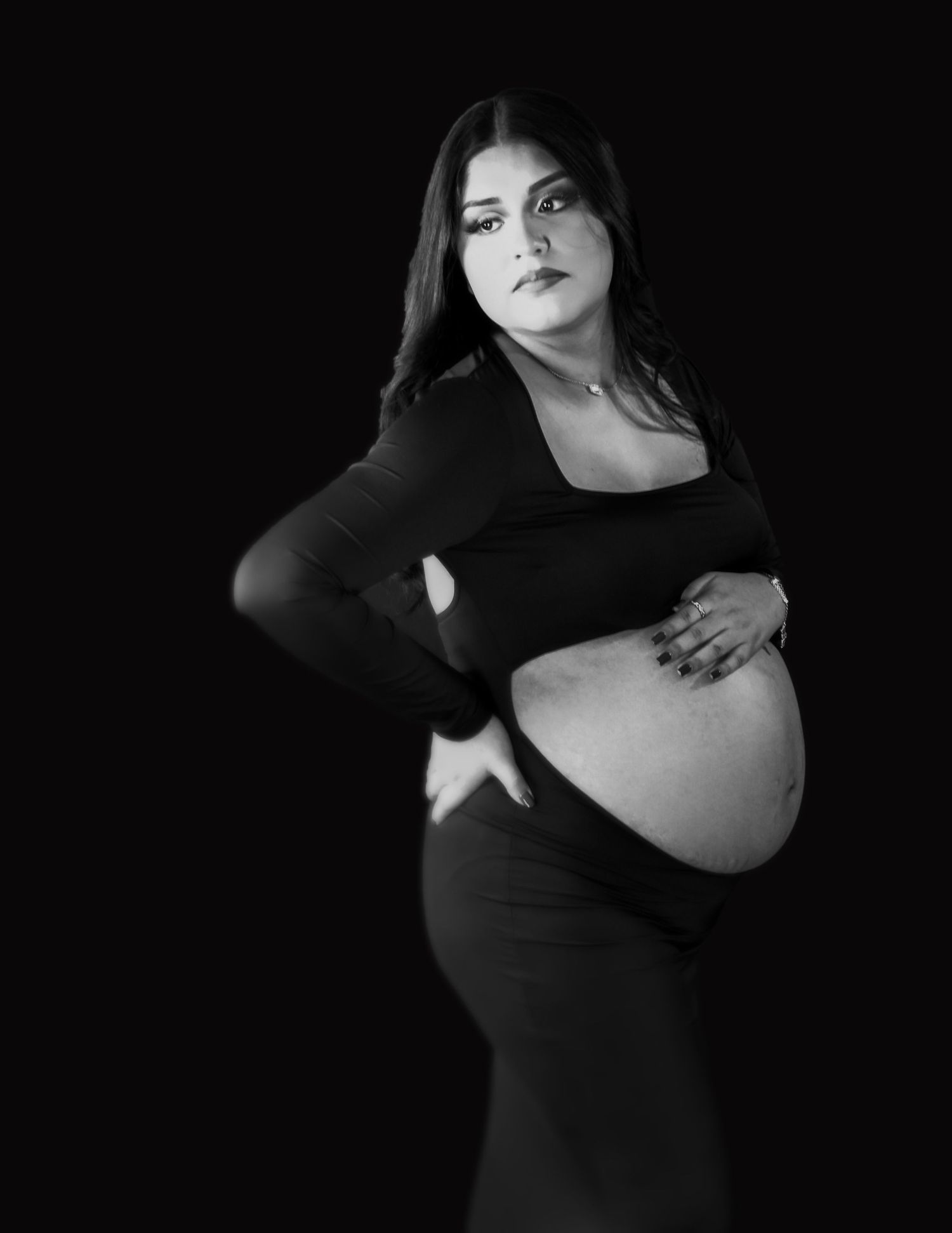A luxury black and white maternity photo showing a beautiful pregnant woman in a dark dress against a dark background holding her belly.