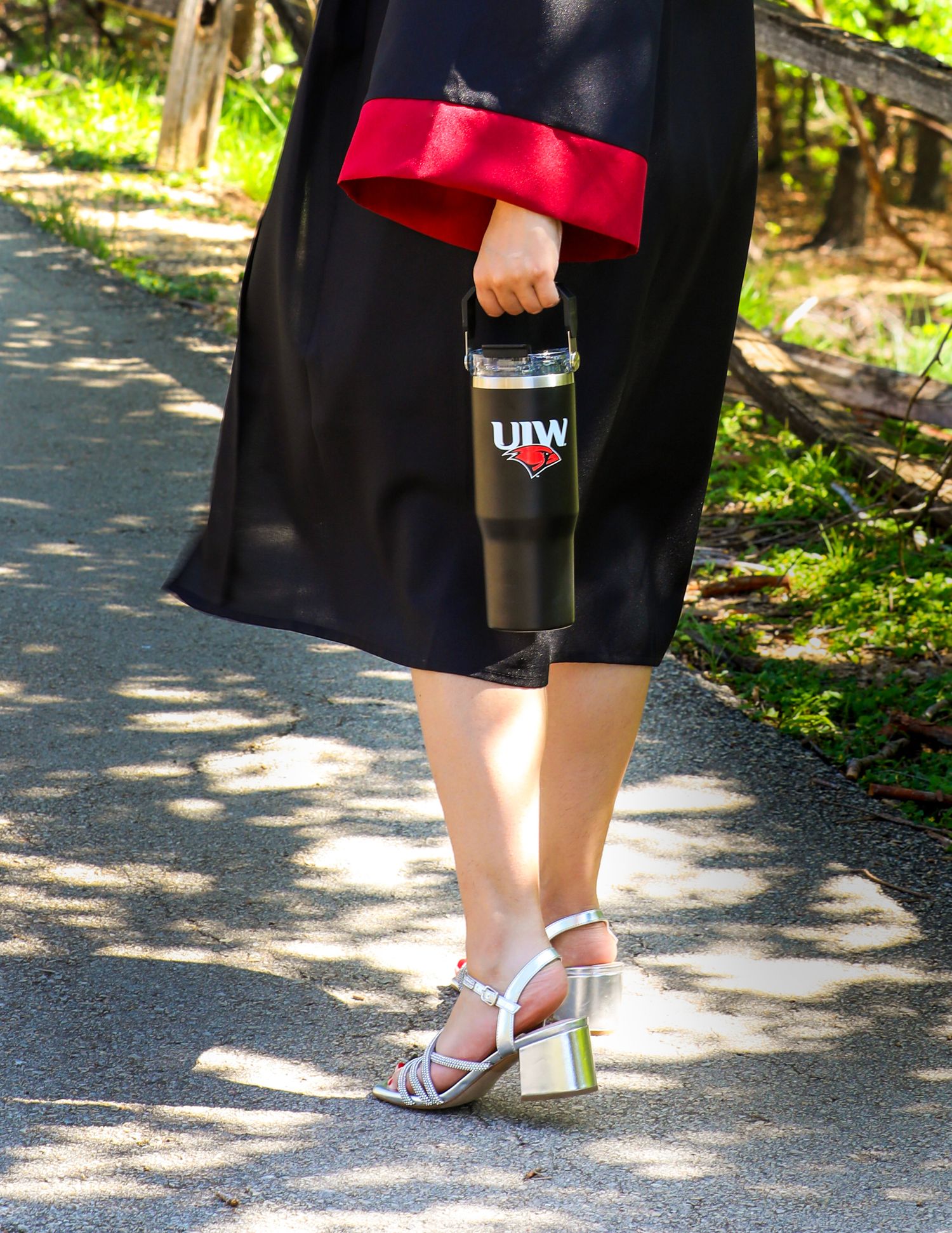 Student wearing cap and gown with red trim and strappy metallic heels holds water bottle posing at Denman Park during her senior photos. UIW