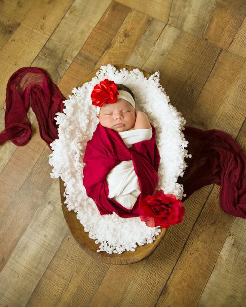 Newborn baby girl swaddled in red wrap with floral headband, sleeping peacefully in a white ruffled basket on wood floor.