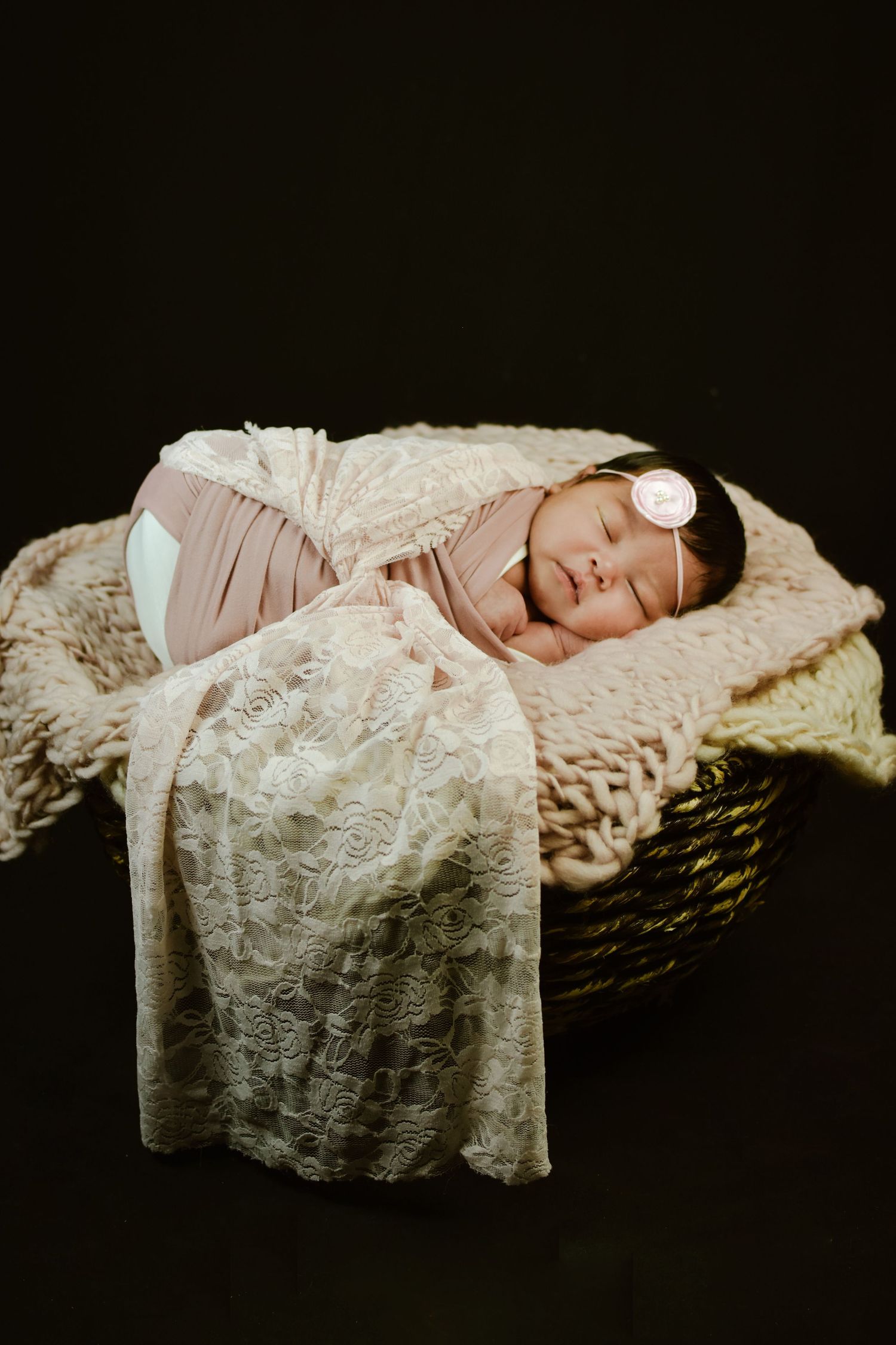 Newborn baby girl swaddled in pink wrap sleeping peacefully in a wicker basket with lace blanket and floral headband.