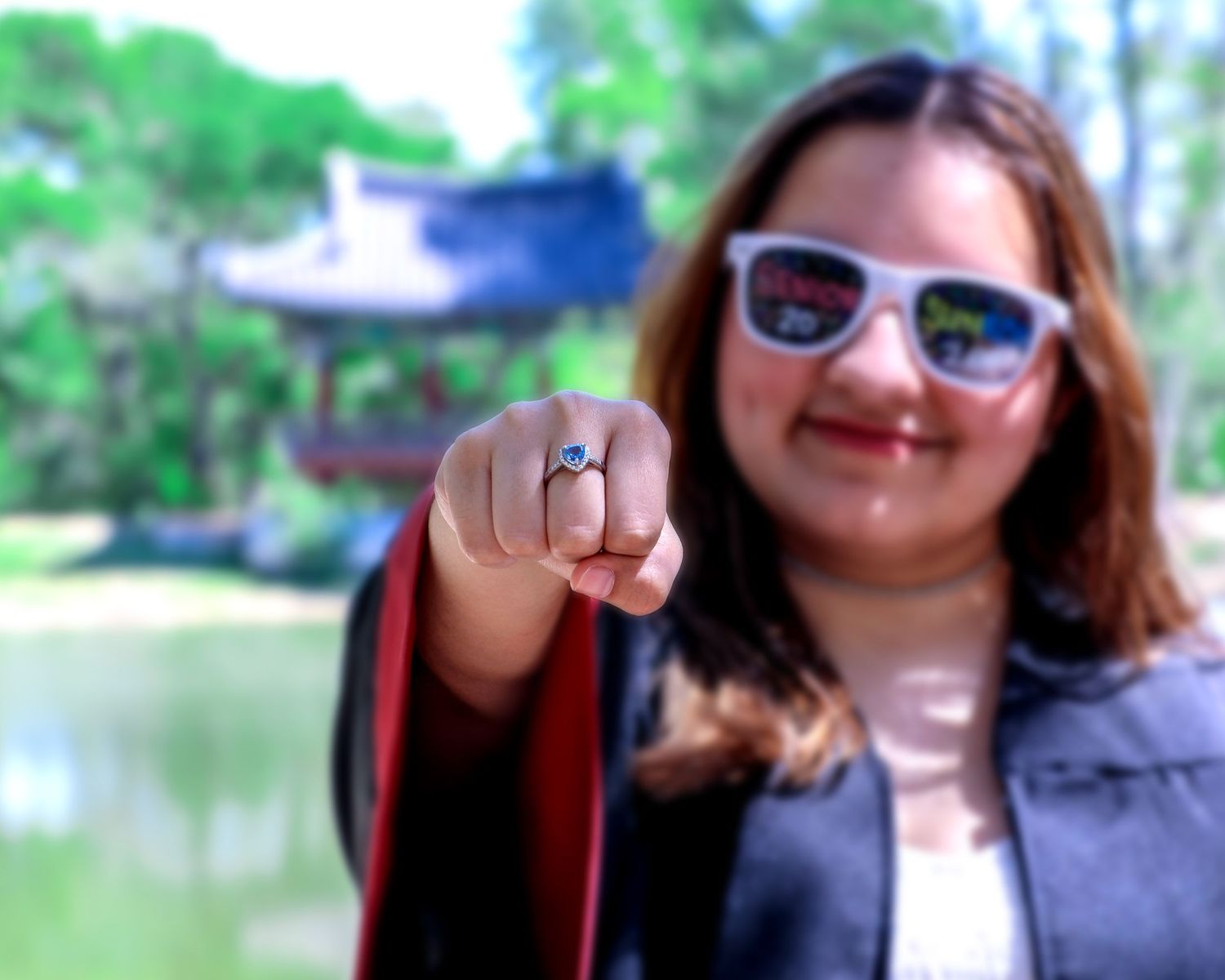 Woman in sunglasses showing off an engagement ring outdoors near a pond with a pavilion in the background.