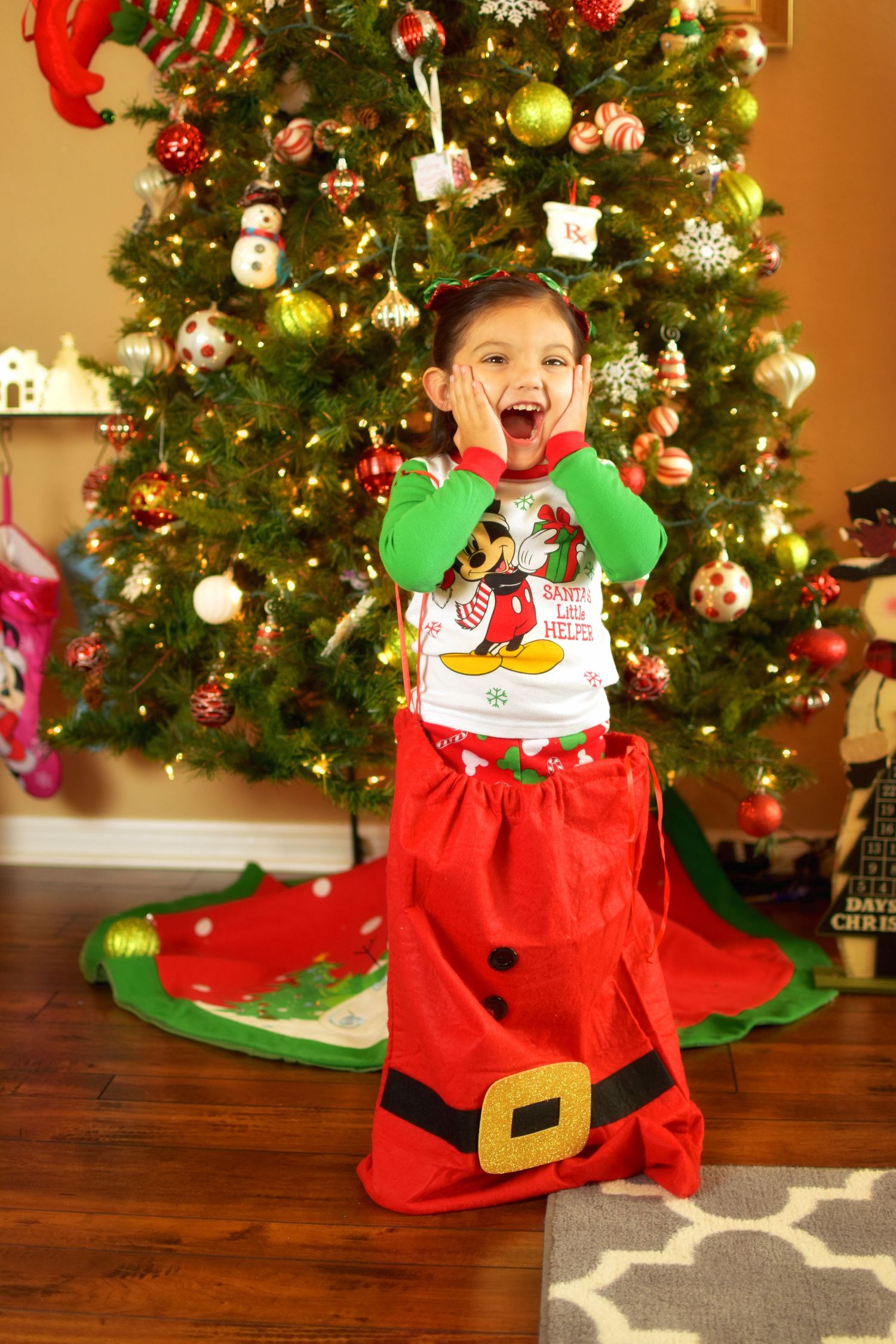 Excited toddler girl standing in a red Santa sack in front of a decorated Christmas tree, wearing Mickey Mouse pajamas.
