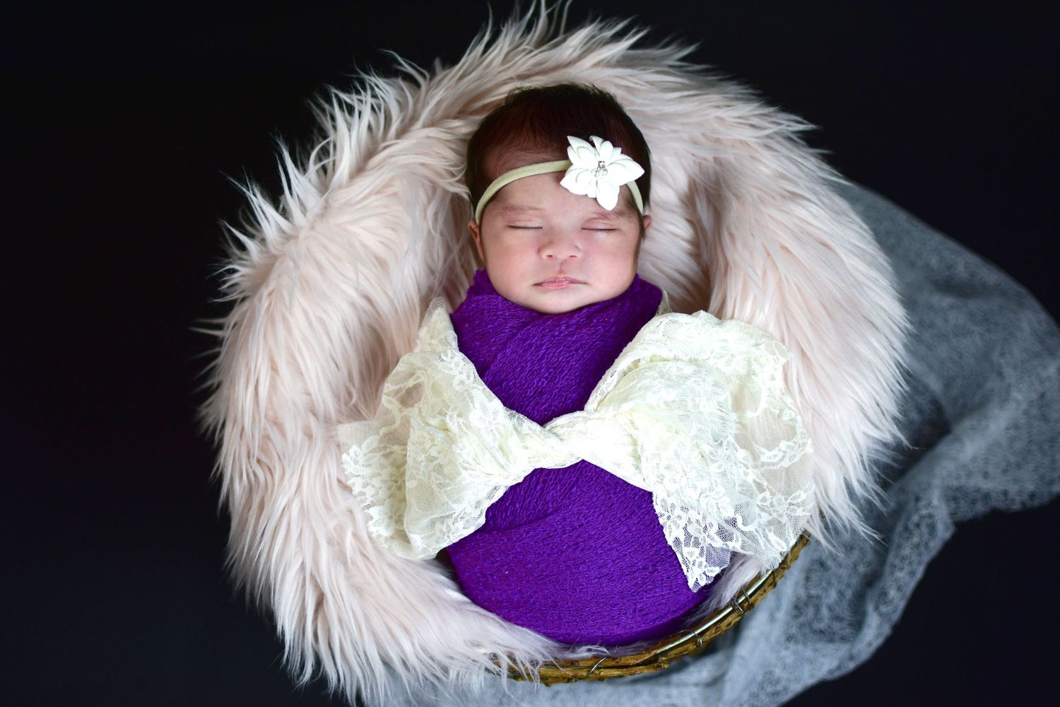 Newborn baby sleeping in a basket wrapped in purple and cream lace, wearing a white flower headband.