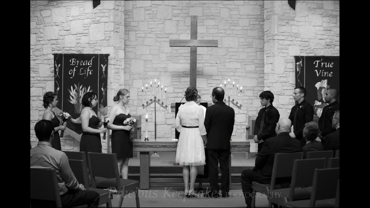Black and white photo of a wedding ceremony at a church altar with cross, bridal party, and guests in pews.