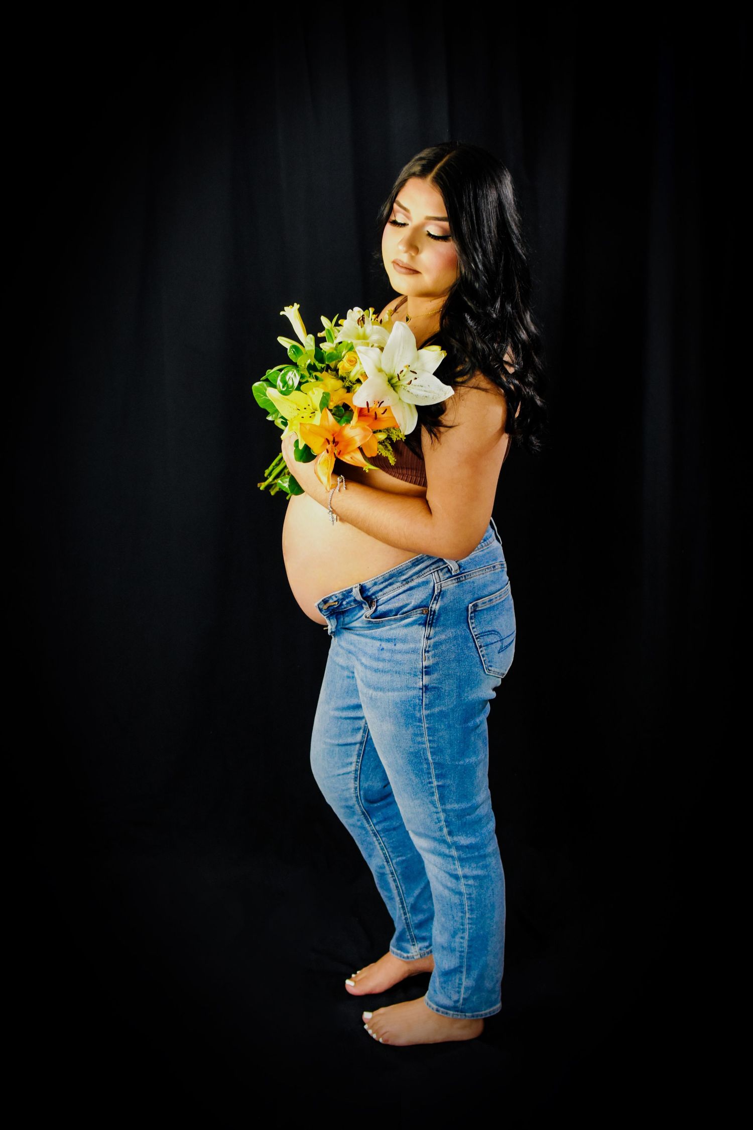 Pregnant Woman in denim poses against dark background holding colorful floral bouquet at her maternity photo session in San Antonio