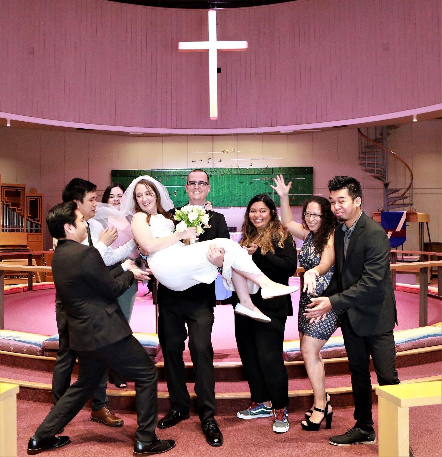 Wedding party celebrating in a pink church, groom carrying bride in white dress, surrounded by joyful guests near a cross.