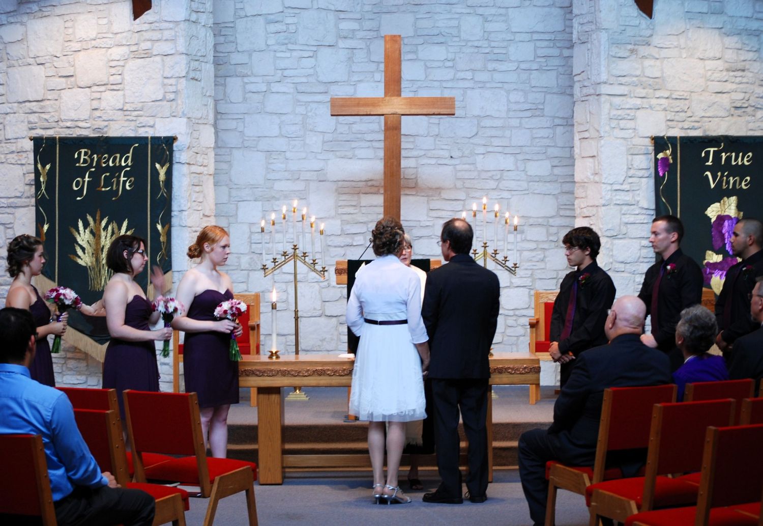 Wedding ceremony in a church with bridesmaids, groomsmen, and couple at the altar beneath a wooden cross.