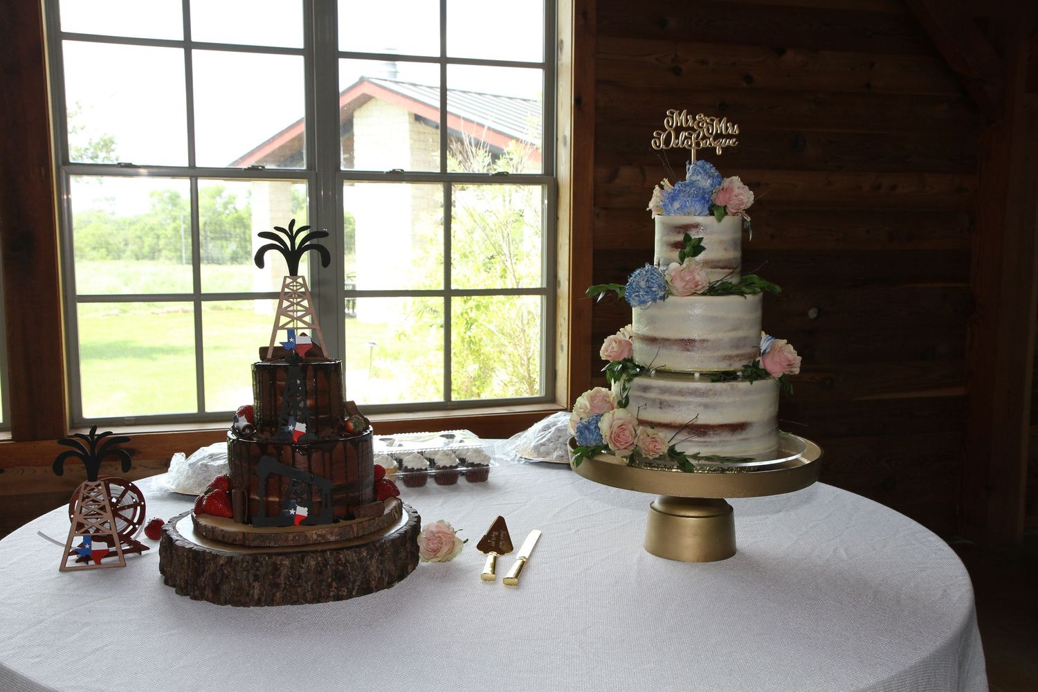 Two elegant wedding cakes displayed on a white table, one dark chocolate and one semi-naked white cake on a gold stand.