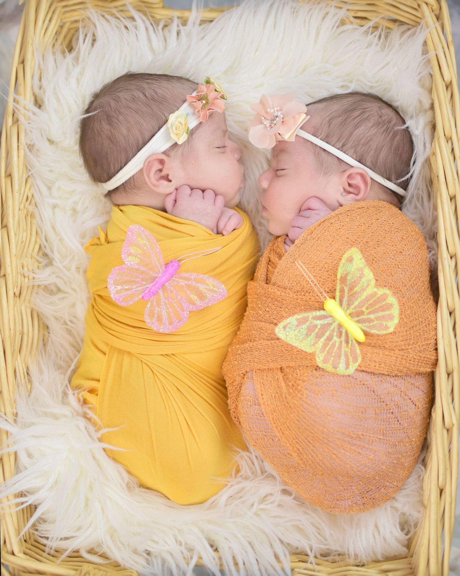Two sleeping newborn twins swaddled in yellow and orange wraps with butterfly decorations and floral headbands in a basket.