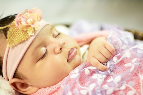 Peaceful newborn baby girl sleeping in a basket, wearing a pink floral headband and wrapped in a purple blanket.